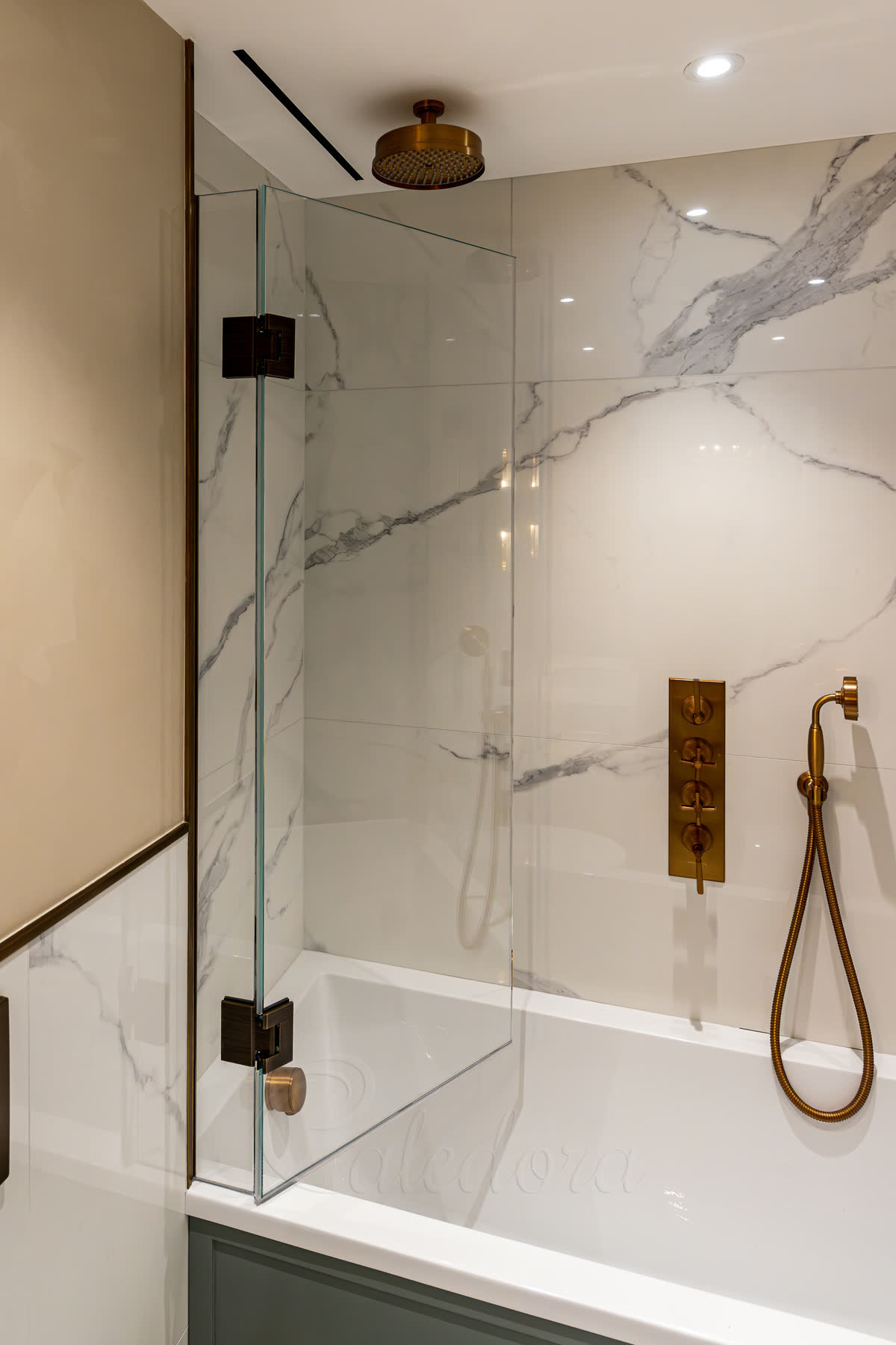 Frameless bath screen with dark antique brass hinges — a fixed glass panel hinged at the wall over the bath, with brass shower head, controls and handset against Calacatta-style marble walls