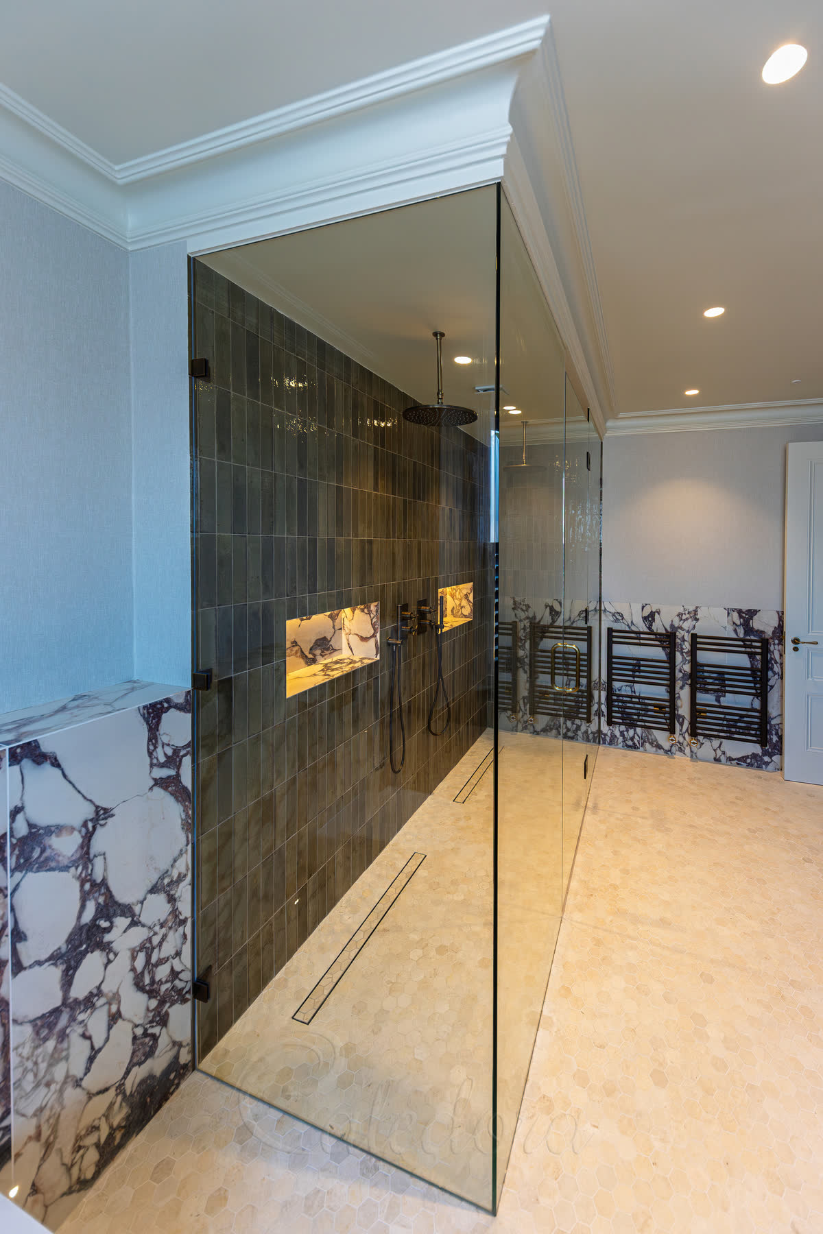 Large brass shower enclosure with brass shower head, controls and handset against deep green marble walls and patterned floor tiles