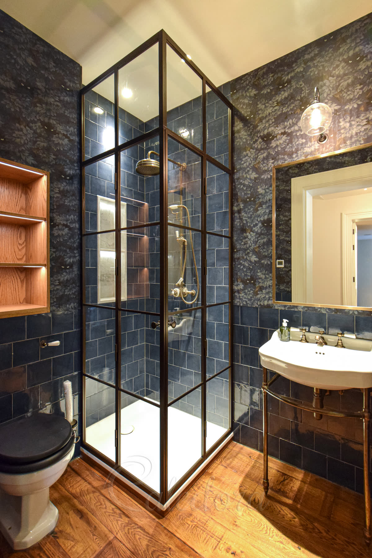Bronze-framed corner shower enclosure with antique brass shower head, controls and handset against a dark bronze grid frame, deep blue patterned wallpaper and dark blue metro tiles inside
