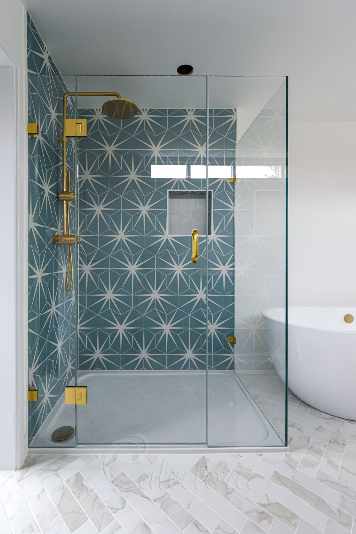 Brushed brass shower fittings — bright satin brass shower head, handle and hinges on a frameless corner shower enclosure against star-pattern teal cement tiles, with a freestanding bath alongside