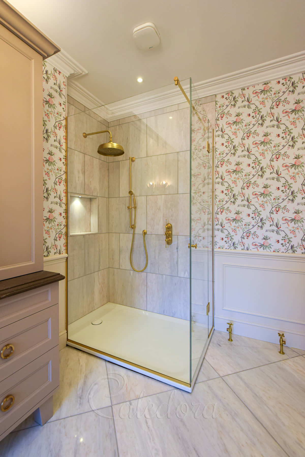 Brushed brass framed shower enclosure with brushed brass top profile and matching shower fittings against marble walls and floral wallpaper