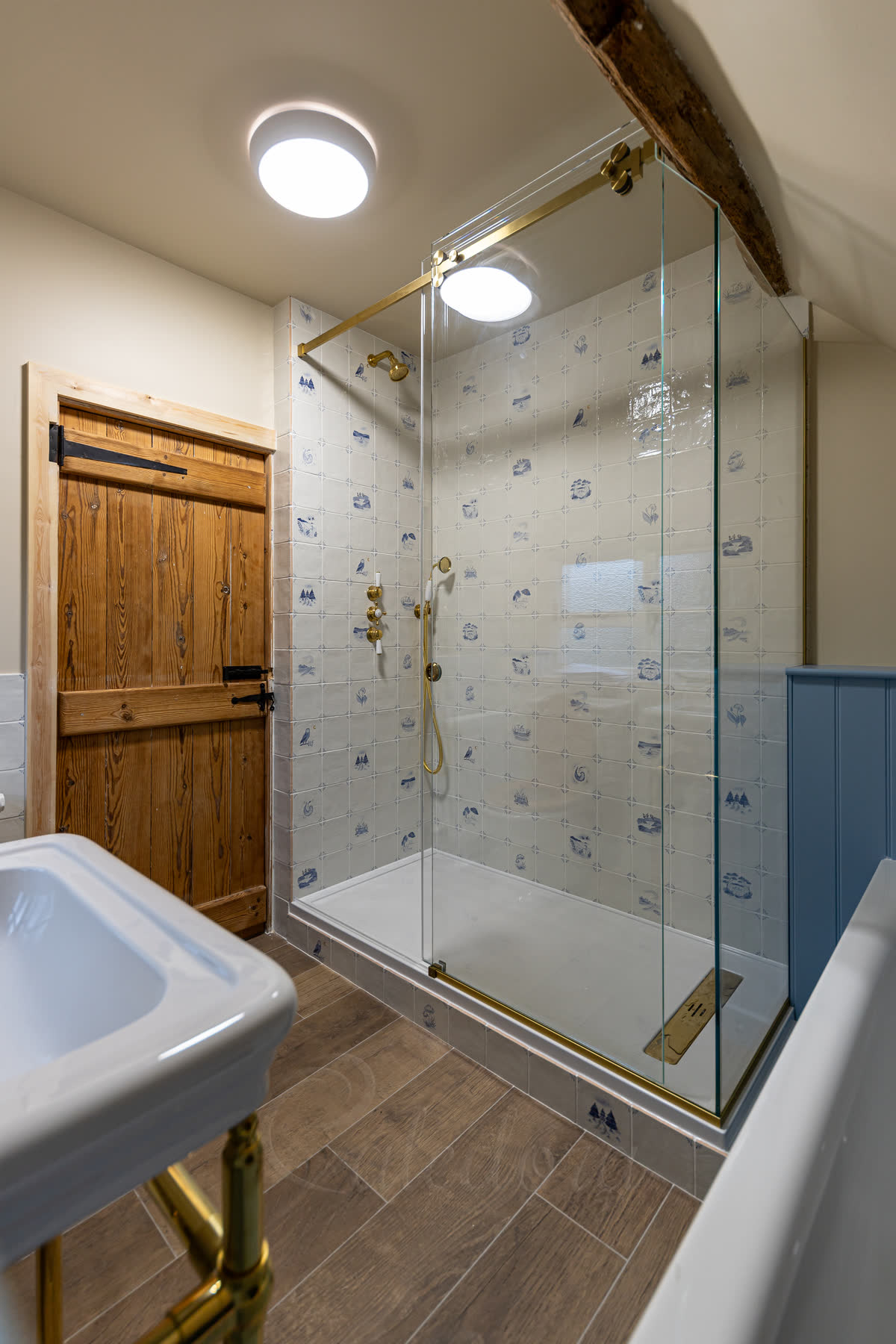 Brushed brass sliding shower door in a loft bathroom — brushed brass sliding track across the top, with matching brushed brass shower head, mixer controls, handset and basin legs against patterned blue-and-white tiles under a sloped ceiling beam