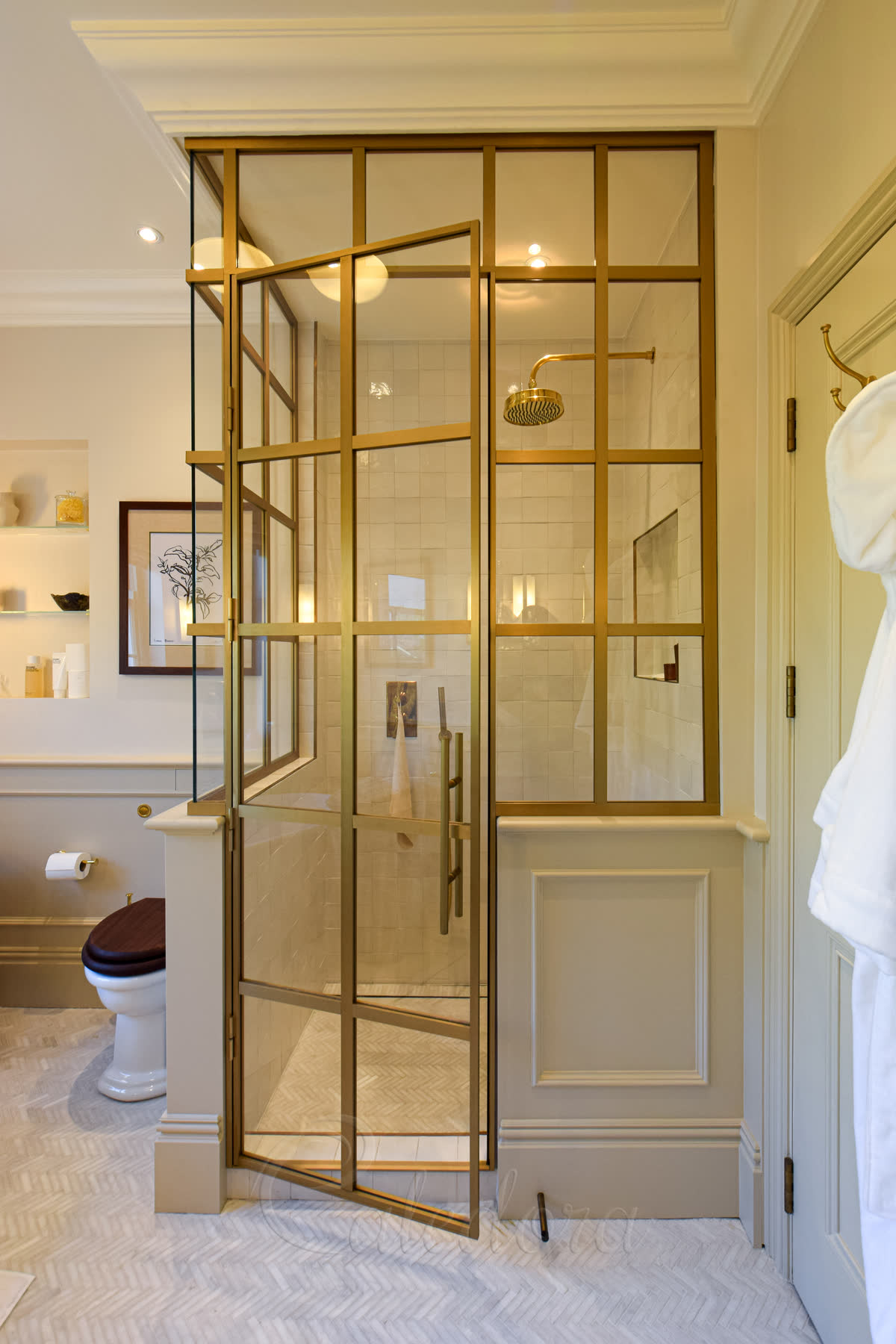 Brushed gold framed shower door open into the enclosure — grid-style brushed gold frame profile with clear glass panels, set against herringbone marble flooring and a panelled half-wall