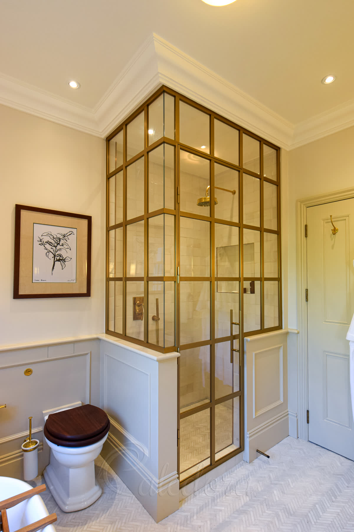 Brushed gold framed corner shower enclosure in a period bathroom — grid-style brushed gold frame around clear glass with a brushed gold shower head inside, panelled cream half-wall, herringbone marble floor and traditional toilet