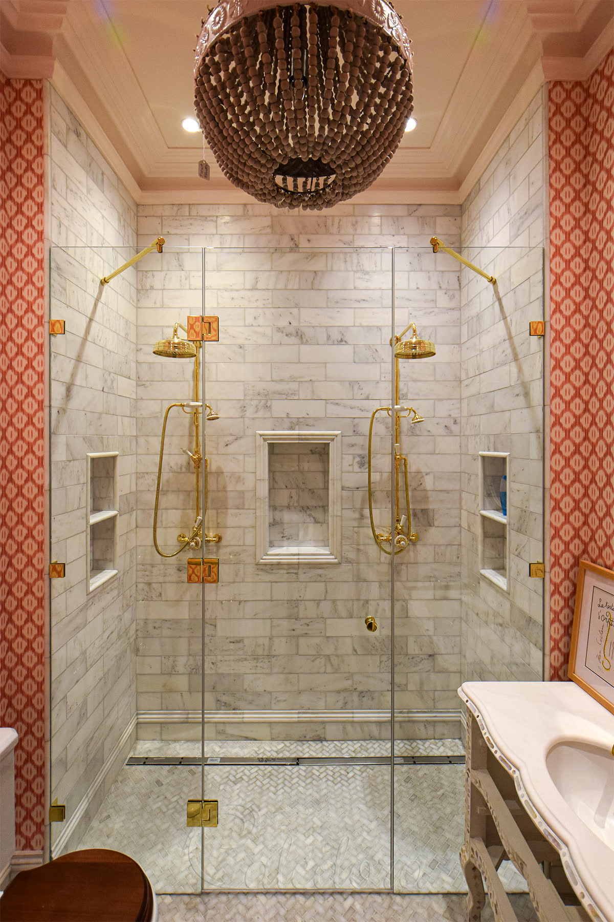 Gold inline shower enclosure recessed between marble walls — two fixed glass panels flanking a central hinged door, with twin gold shower heads, riser rails, handsets and corner brackets against marble metro tiles and a herringbone marble floor