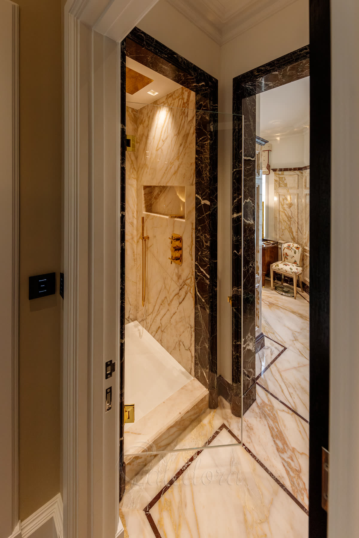 Gold shower door partially open in a marble bathroom suite — bright gold hinges and handle against pale marble walls