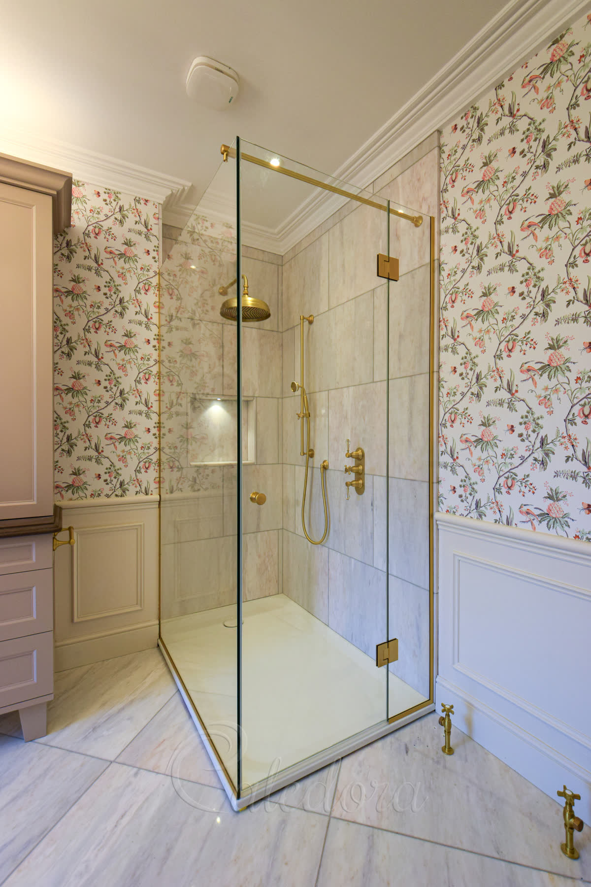 Lacquered brushed-brass corner shower enclosure — brushed brass top profile, hinges and shower fittings preserved by a clear lacquer coating, set against marble walls inside, floral wallpaper outside and a panelled cream half-wall