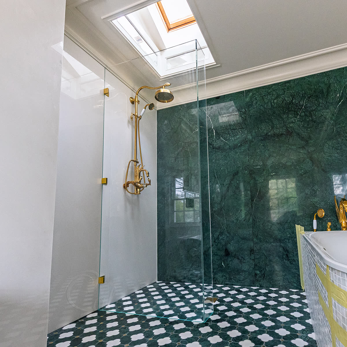 Polished brass shower screen — fixed walk-in panel with polished brass brackets at the wall and floor channel below, set against a green marble wall and patterned green-and-white floor tiles, with matching polished brass shower head, controls and handset