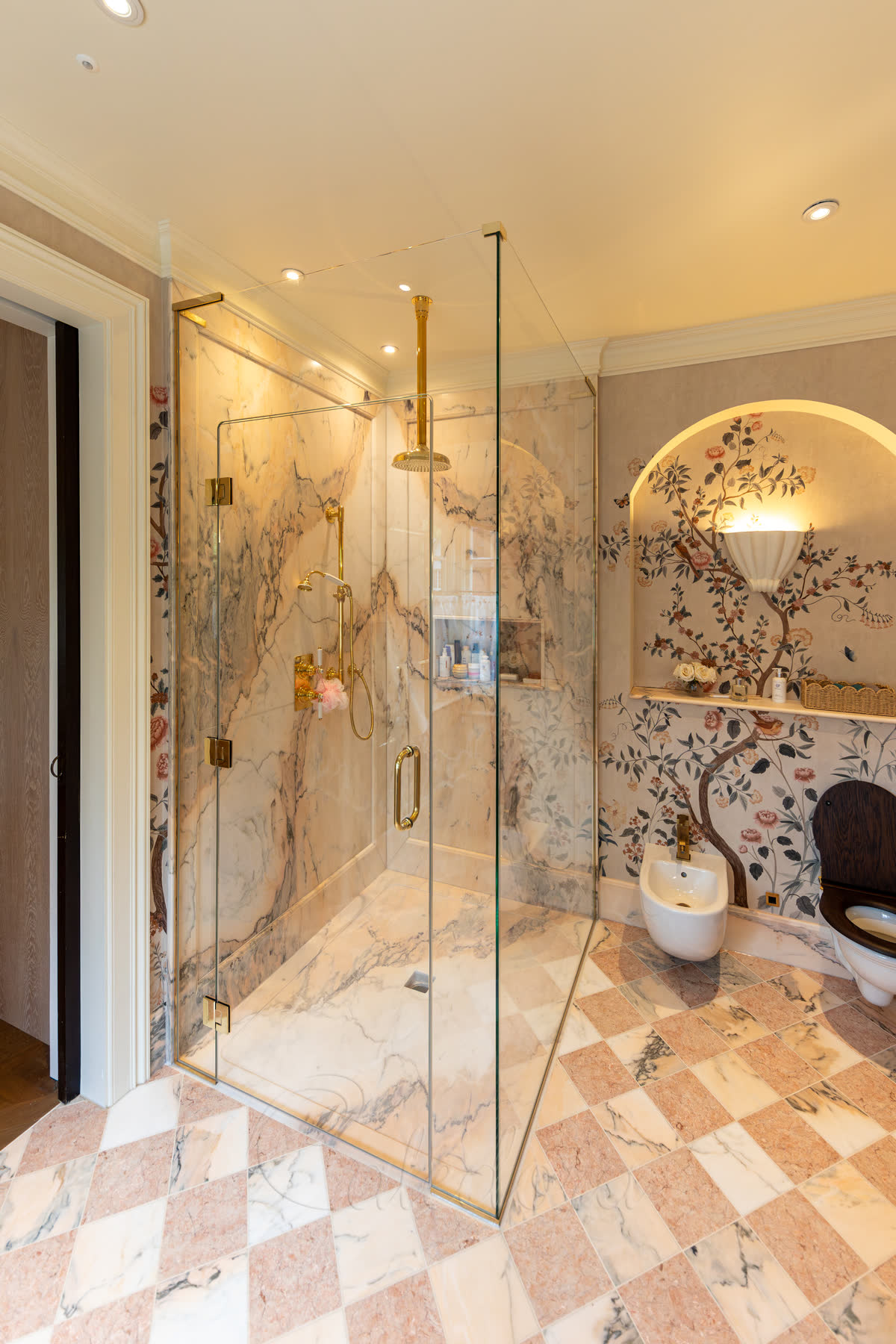 Large unlacquered polished-brass shower enclosure with cut-face door — the front face panel forms a continuous inverted U around an inset door. Unlacquered polished brass hinges, handle, shower head and controls in their natural state, set against Calacatta marble walls inside, a herringbone marble floor and a hand-painted floral mural in an arched alcove