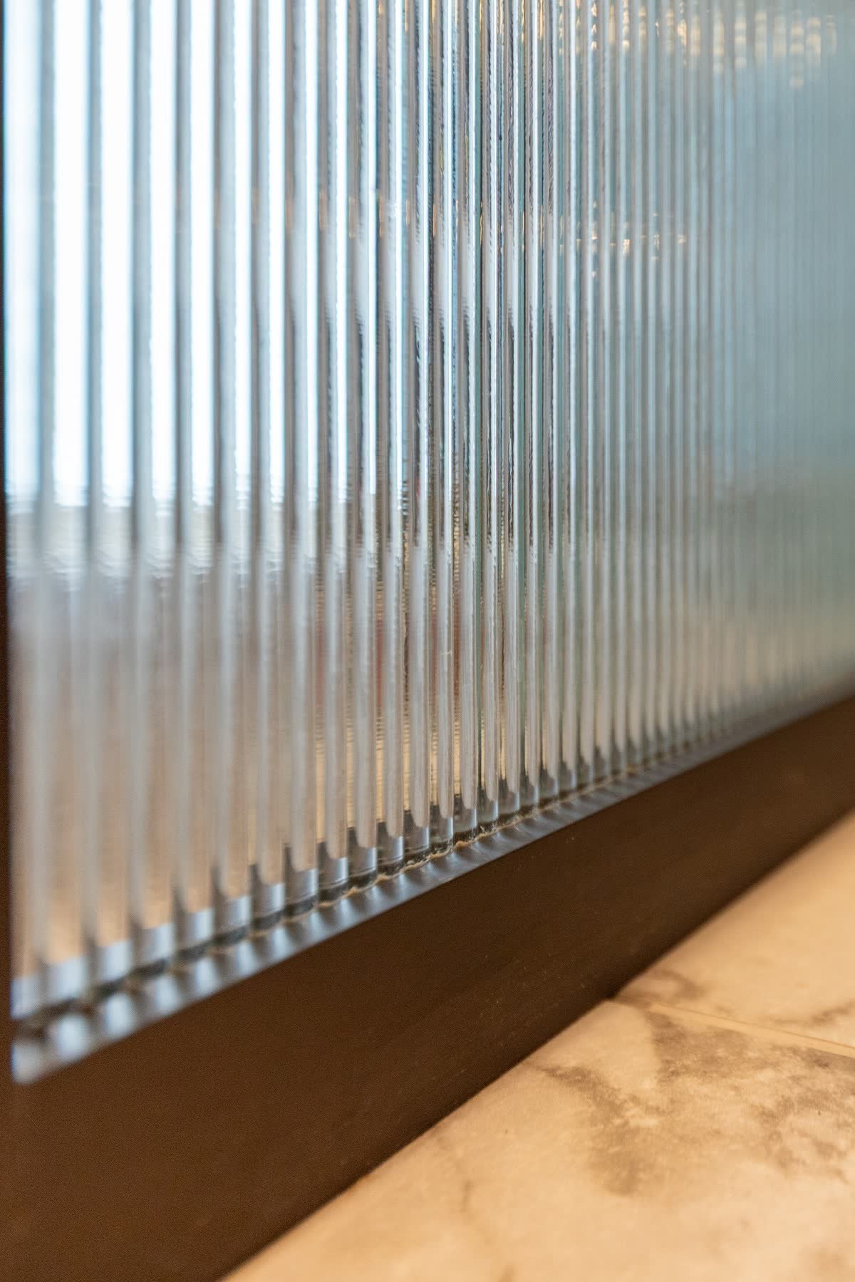 Close view along a fluted glass panel at a raking angle — rounded ribs standing proud of the surface catch the light one after another, their convex profile visible in cross-section where the glass meets the dark frame above a marble sill