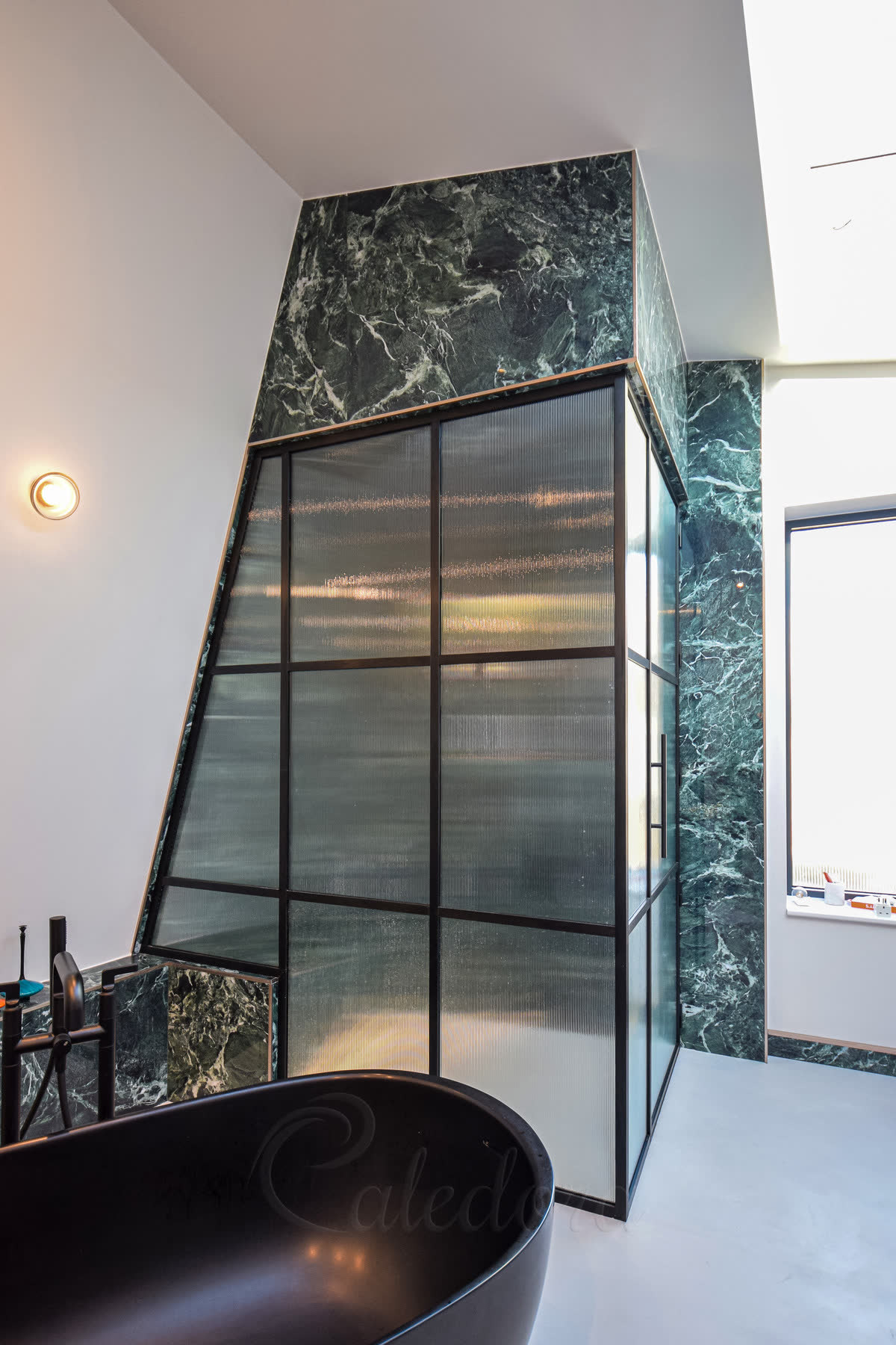 Fluted glass shower panel with its top edge angled to follow a loft roof pitch, in a brushed black grid frame above a black freestanding bath