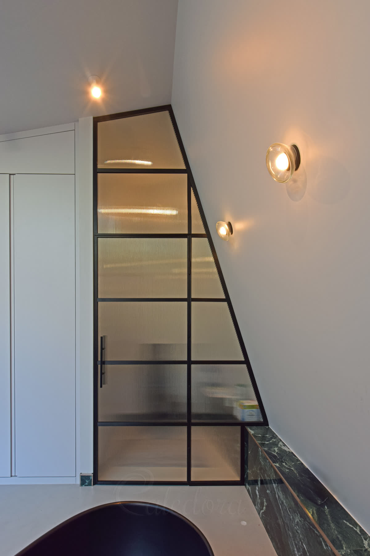 Fluted glass shower partition under a sloped ceiling — black grid framing follows the roof pitch above a black bath, with vertically textured glass panes and dark green marble at the skirting