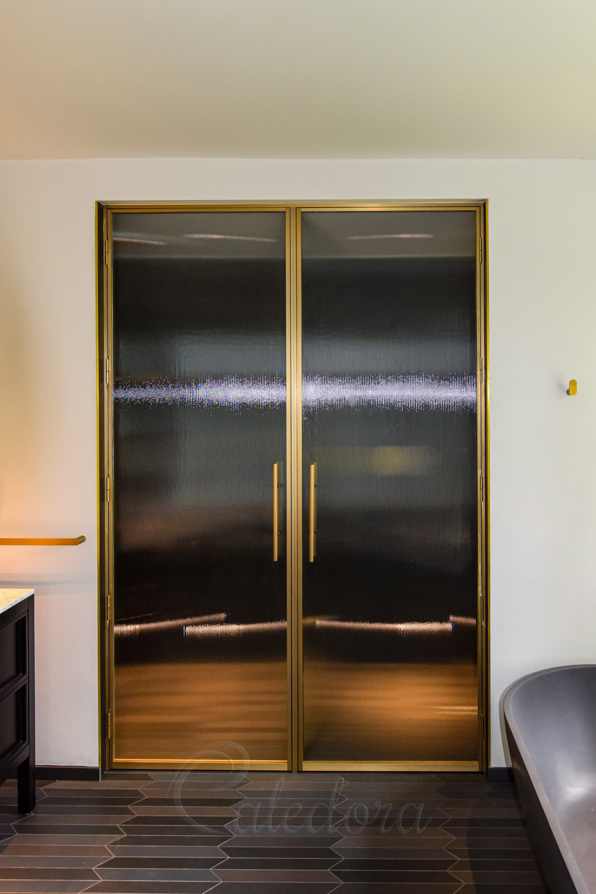 Fluted glass shower doors in a brushed brass frame — a pair of full-height double doors glazed in fluted laminated glass, slim brushed brass framing and matching bar handles, set into a white wall above a dark hexagonal tiled floor