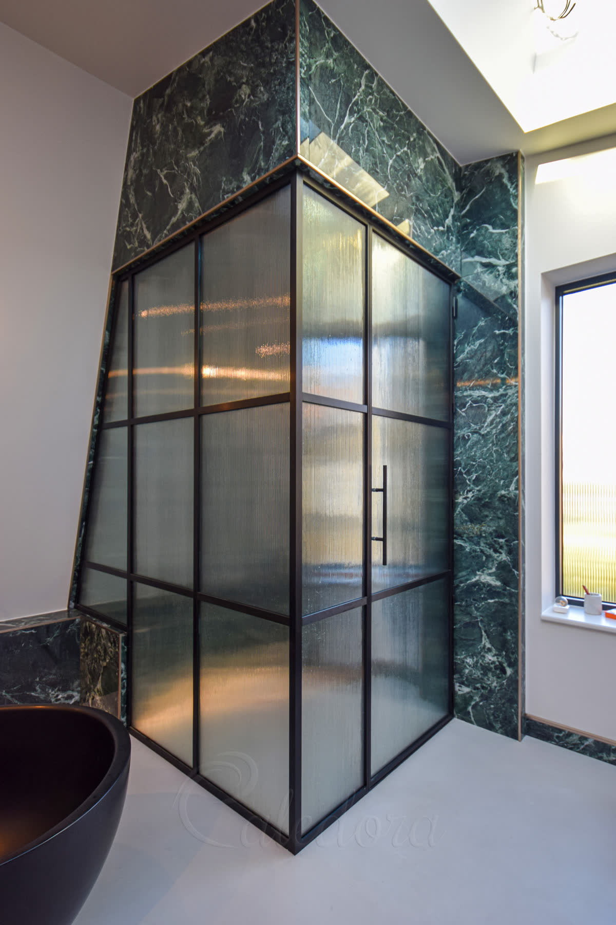 Fluted glass shower enclosure in a brushed black grid frame — a corner enclosure glazed in 6 mm fluted glass and divided into panes by slim black bars, one side following a sloped loft ceiling, against dark green marble walls