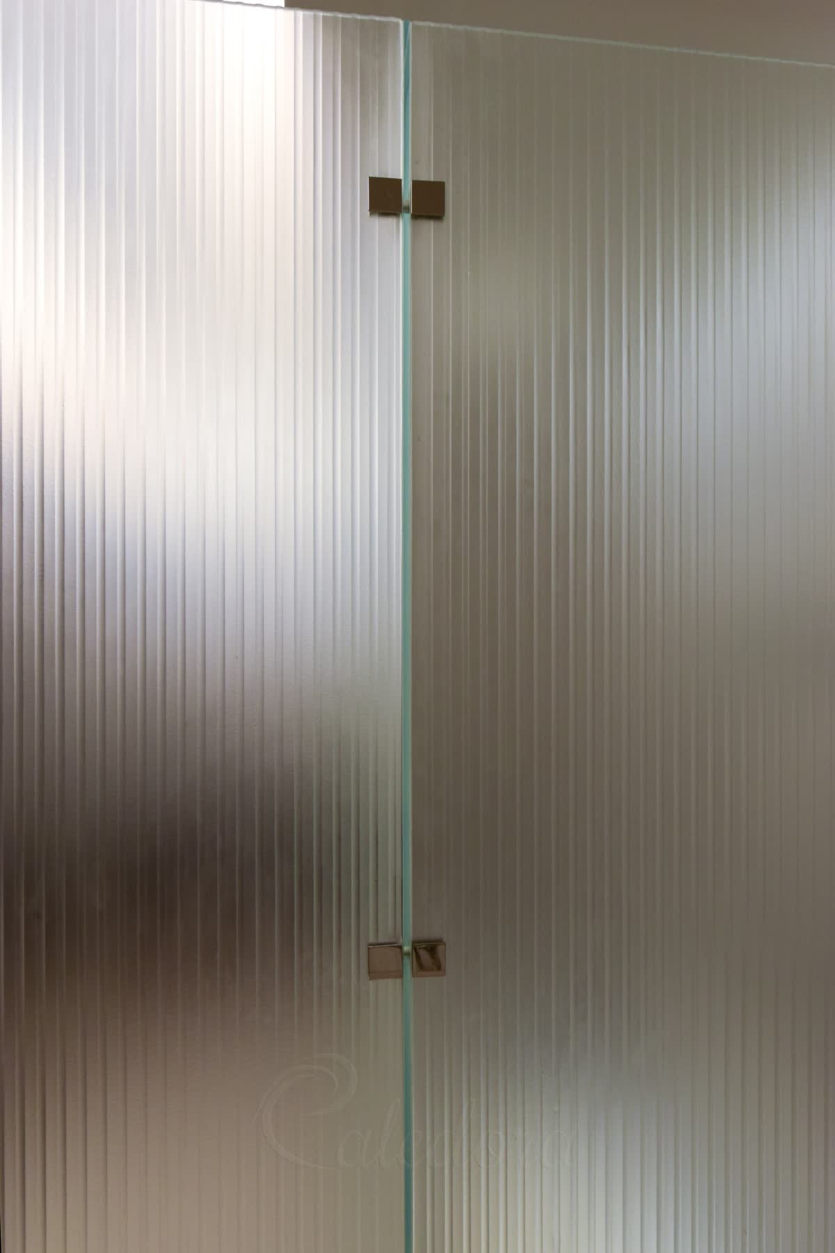 Close view of kiln-formed glass that has been sandblasted — two panels meeting at a polished nickel bracket, the vertical ribs softened to a matt finish that hides what is behind them