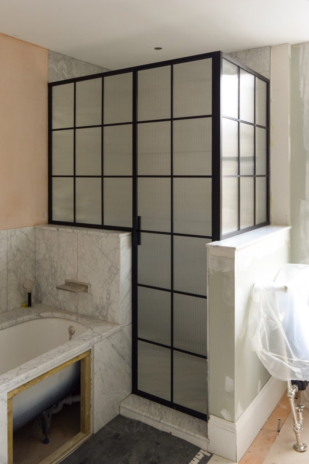Reeded glass enclosure in a black grid frame built over a bath, with a Carrara marble surround