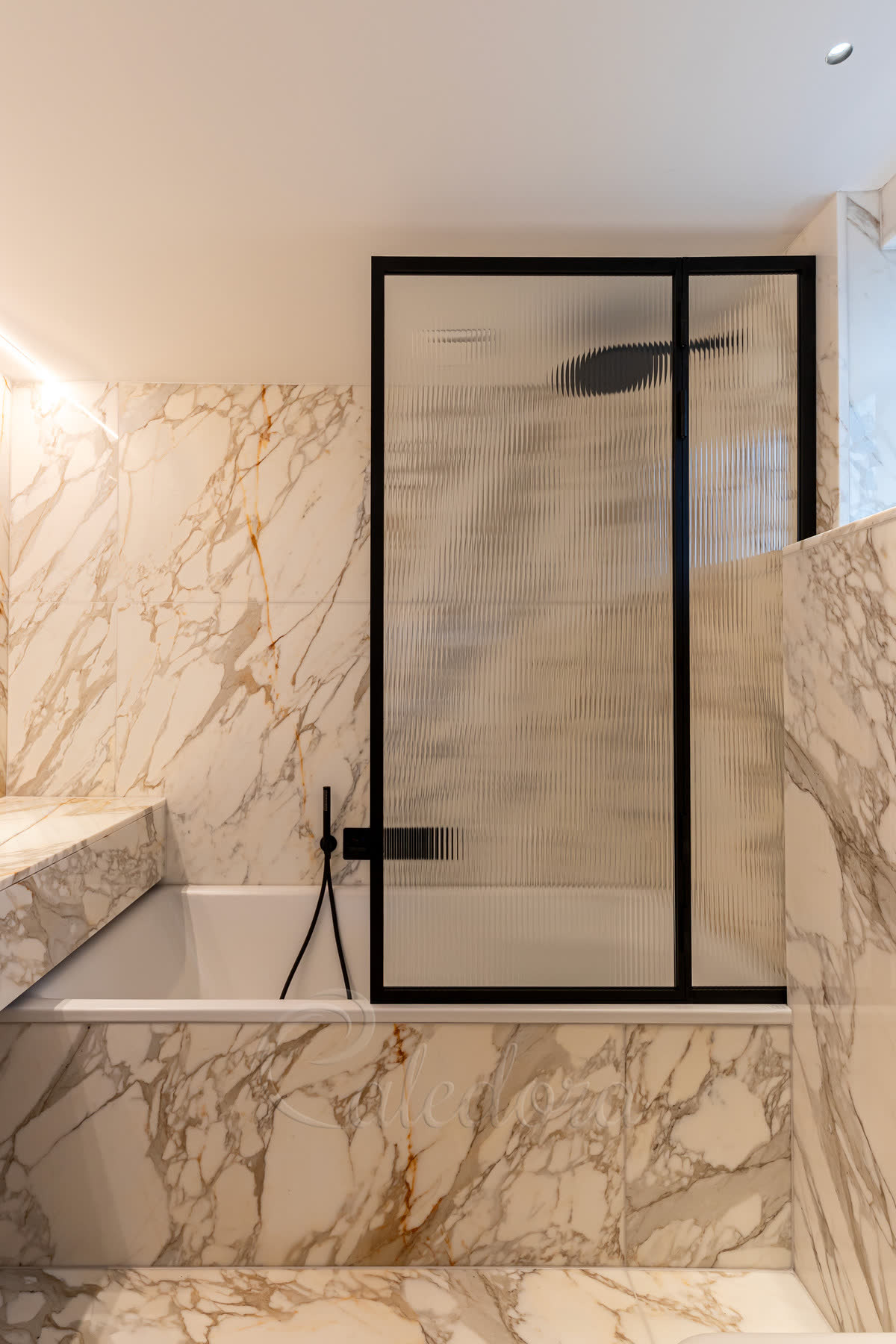 Reeded glass bath screen in a black frame folding over the end of a bath, against Calacatta marble