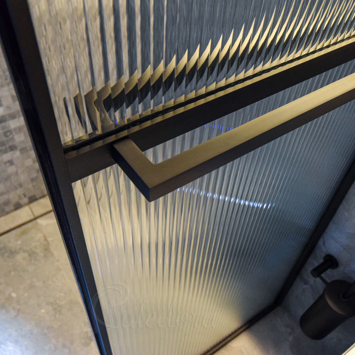 Reeded glass in a slim black frame — close view of the frame junction and matching black bar handle laid across the vertical ribs, the glass edge catching the light where two panels meet