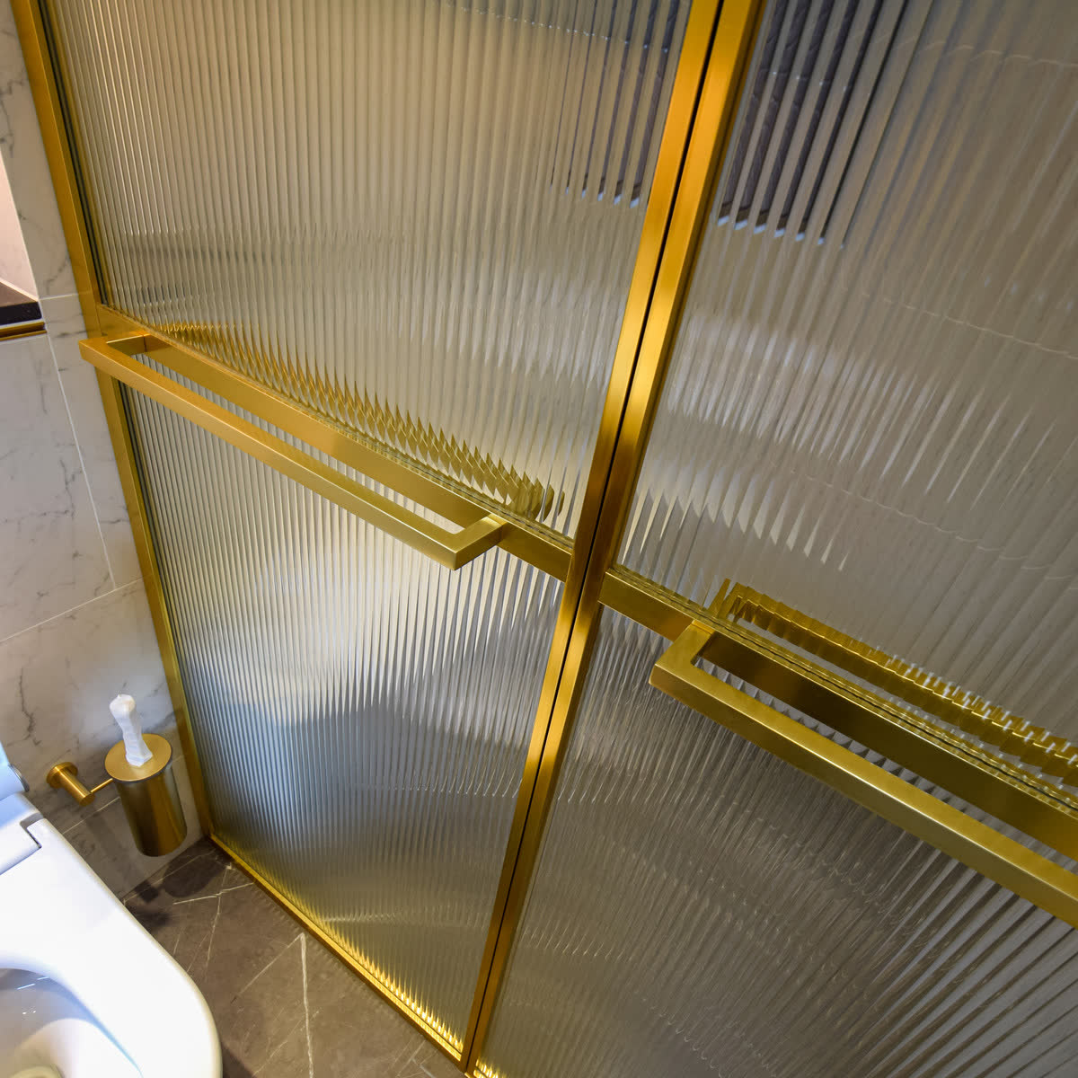 Reeded glass in a brushed brass frame — close view of the frame junction and matching brass bar handles crossing the vertical ribs, against marble