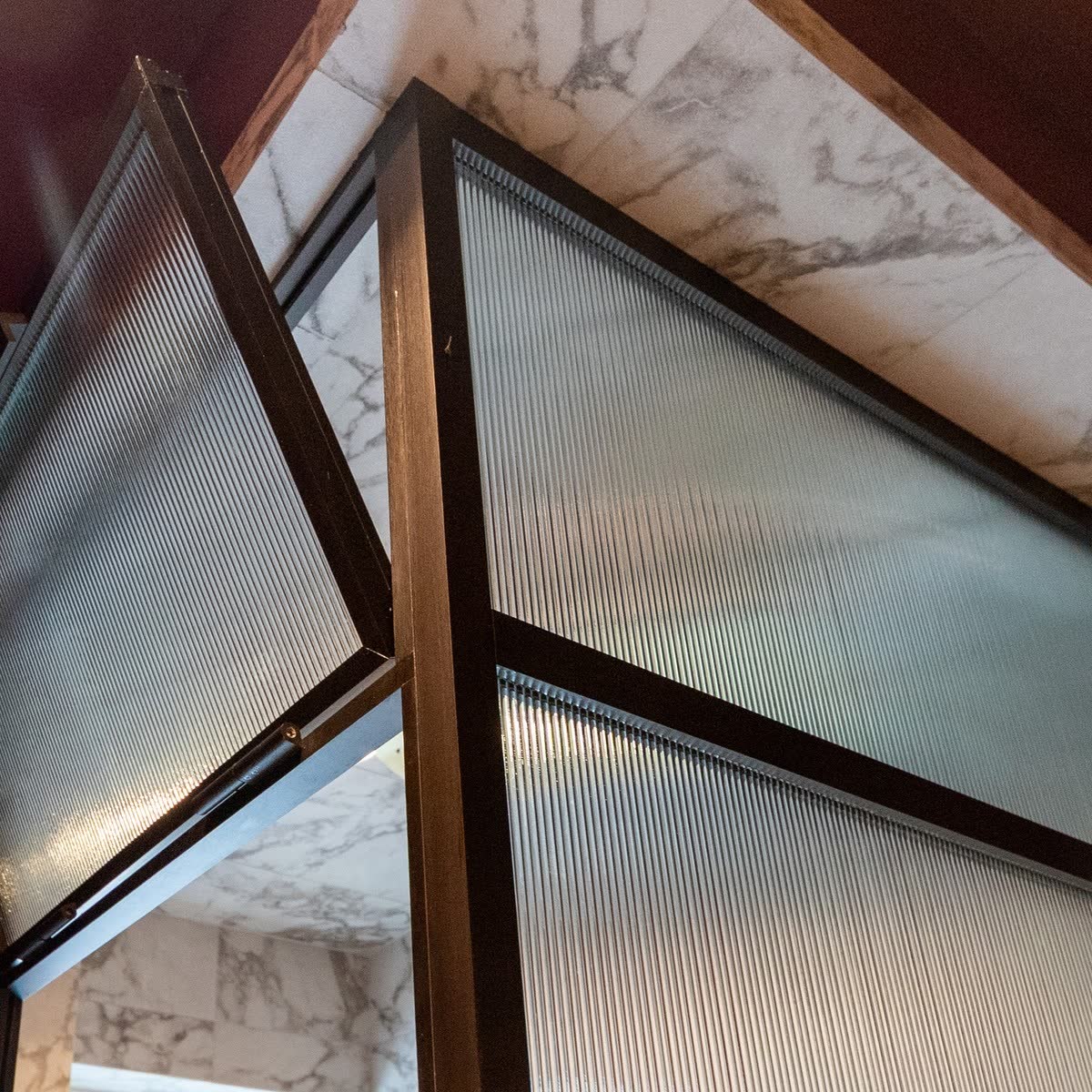 Reeded glass in a dark bronze frame — close view of the frame members meeting at the head of an enclosure, the warm dark metal reading noticeably deeper than brass against marble and a burgundy ceiling
