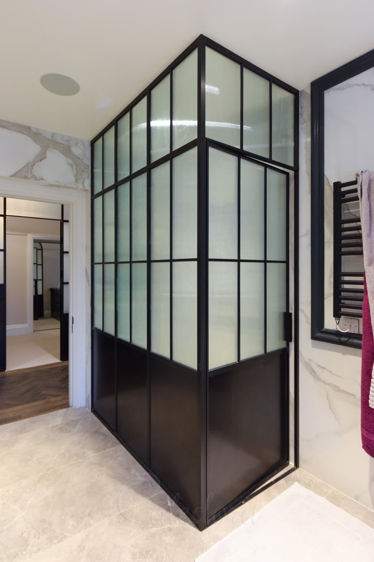 Reeded glass corner shower enclosure in a black grid frame with solid dark lower panels, against Calacatta marble with a black towel radiator