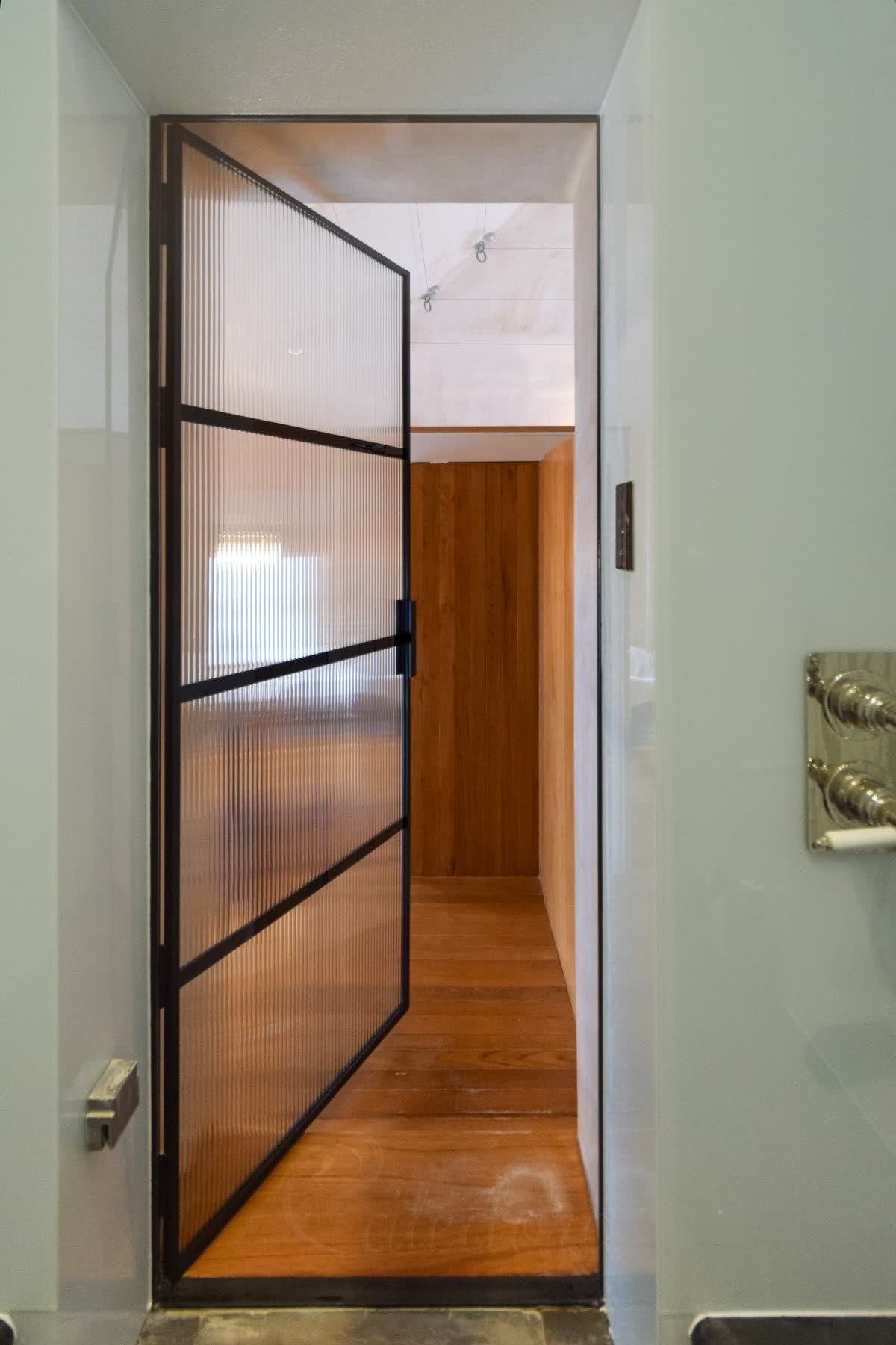 Black framed reeded glass door standing open, seen from inside a shower with pale green glass walls, looking out to an oak-lined room and timber floor
