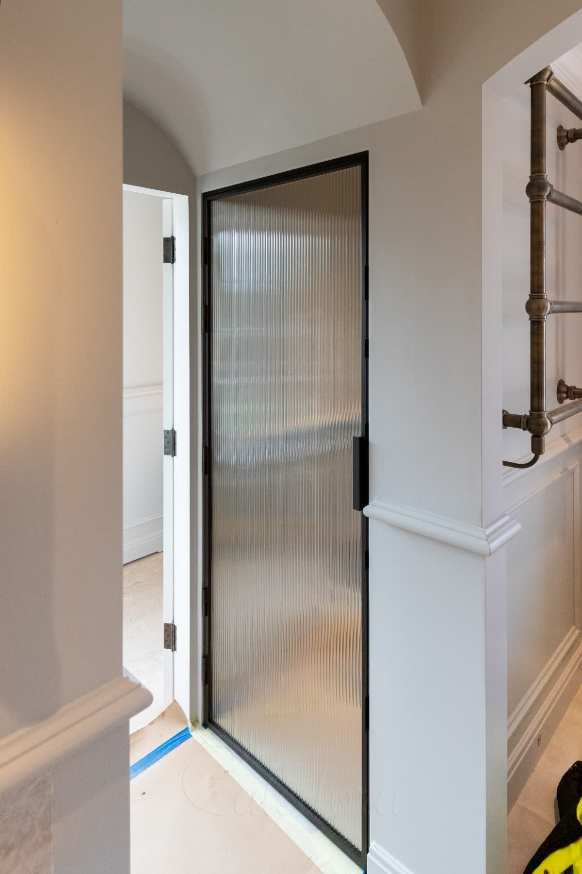 Reeded glass door in a black frame set into a panelled corridor beneath a barrel-vaulted ceiling, beside an antique brass towel rail