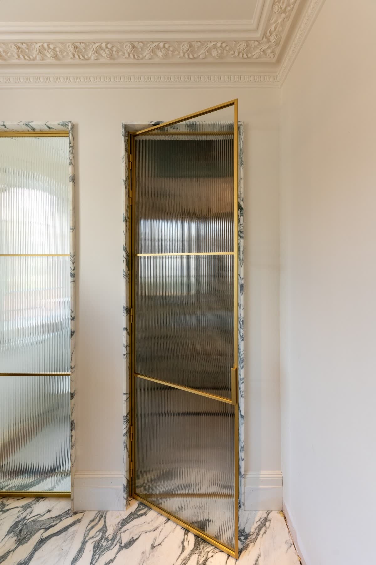 Reeded glass door in a slim brass frame standing open in a marble-lined opening, beneath an ornate plaster cornice with Arabescato marble reveals and floor