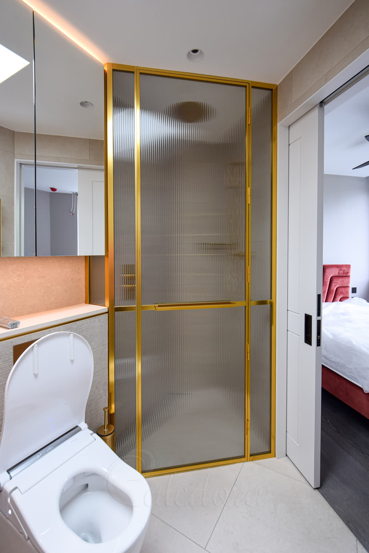 Reeded glass shower enclosure in a gold frame — a corner enclosure of vertically reeded glass in slim polished gold framing with a matching gold handle, in a bright bathroom opening onto a bedroom beyond