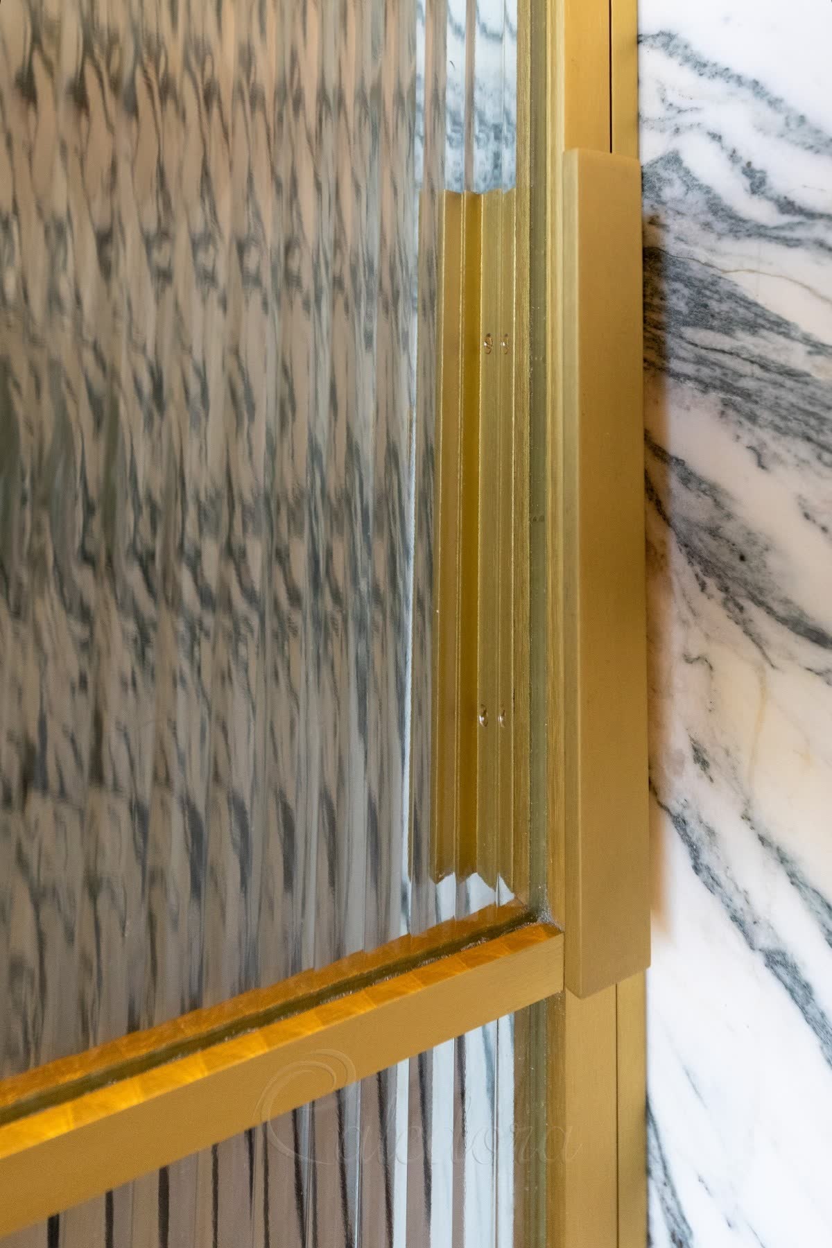 Close view of reeded glass in a brass frame — wide shallow flutes cut into the face rather than raised from it, the scalloped edge visible where the glass meets the brass against veined marble