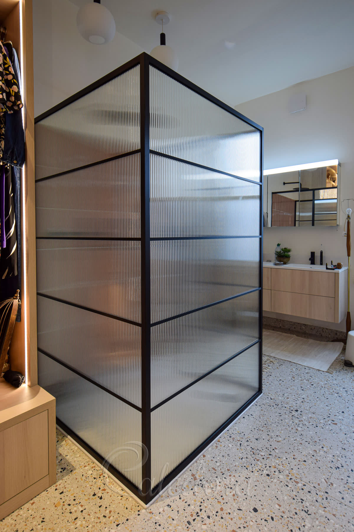 Reeded glass walk-in shower screen in a black grid frame on a terrazzo floor beside pale oak joinery and terracotta tile