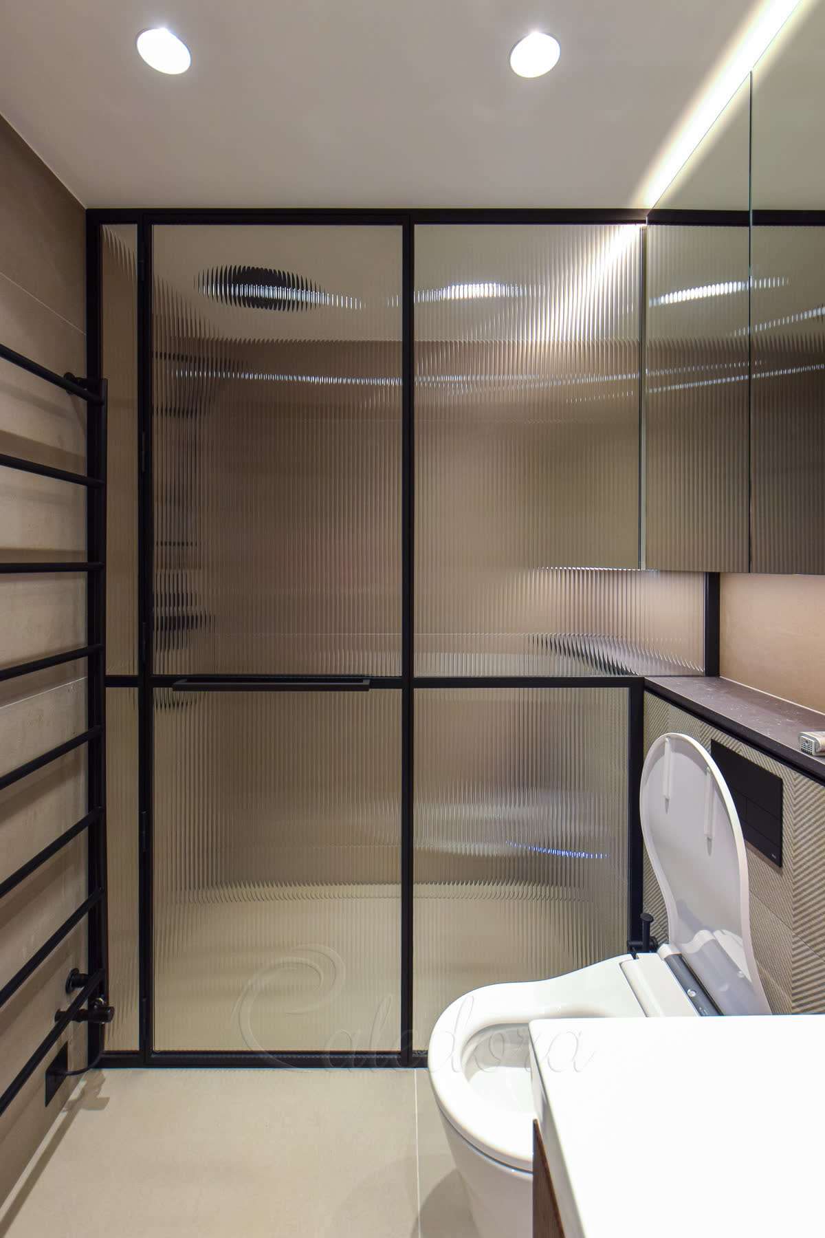 Reeded glass shower enclosure in a slim black frame — a corner enclosure of vertically reeded glass with the door closed and a black bar handle, against warm stone-effect walls with a black ladder radiator alongside