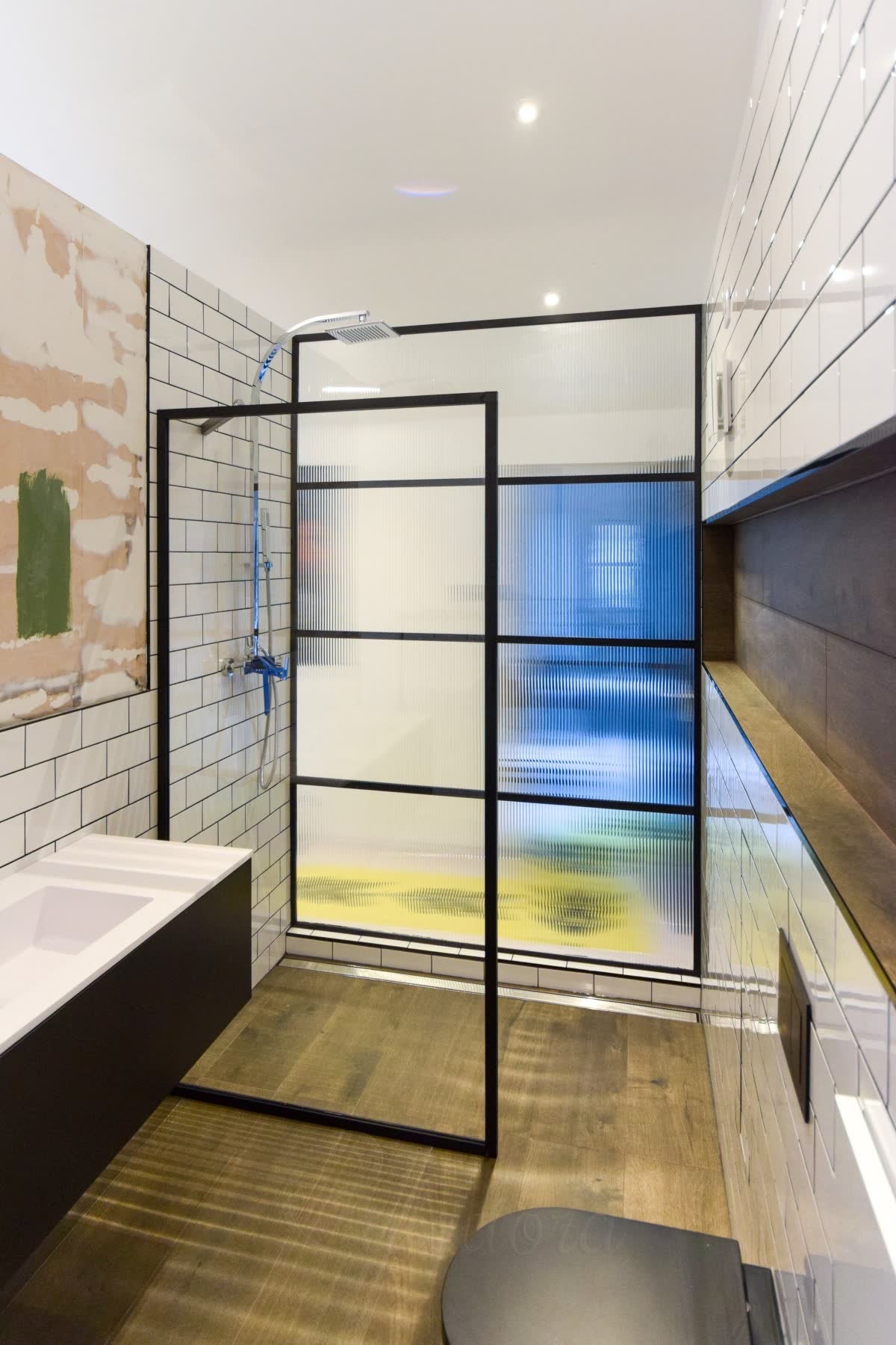 Reeded glass walk-in shower screen in a black grid frame against white metro tile, with colour from the room beyond breaking up through the ribs