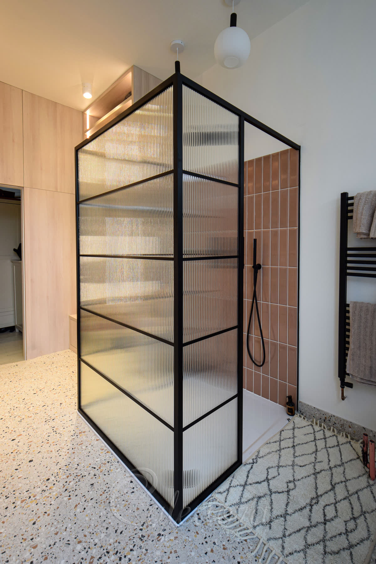 Reeded glass walk-in shower screen — a freestanding black grid screen wrapping two sides of the shower with the entry left open, terracotta tile and a black handset inside, standing on a terrazzo floor