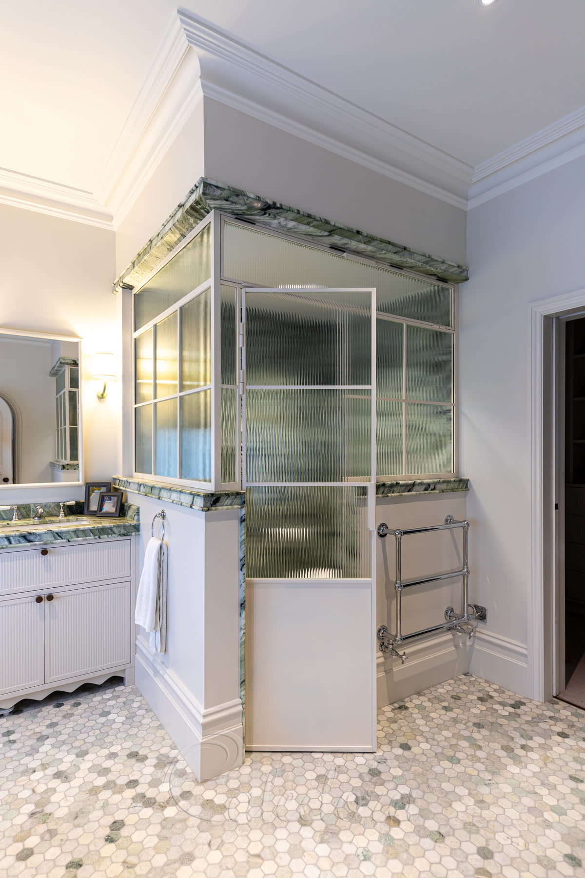 Reeded glass shower enclosure in a white grid frame — a corner enclosure of vertically reeded glass divided into panes by slim white bars, with a green marble top rail and vanity top, a chrome towel rail and a hexagonal mosaic floor in a period bathroom
