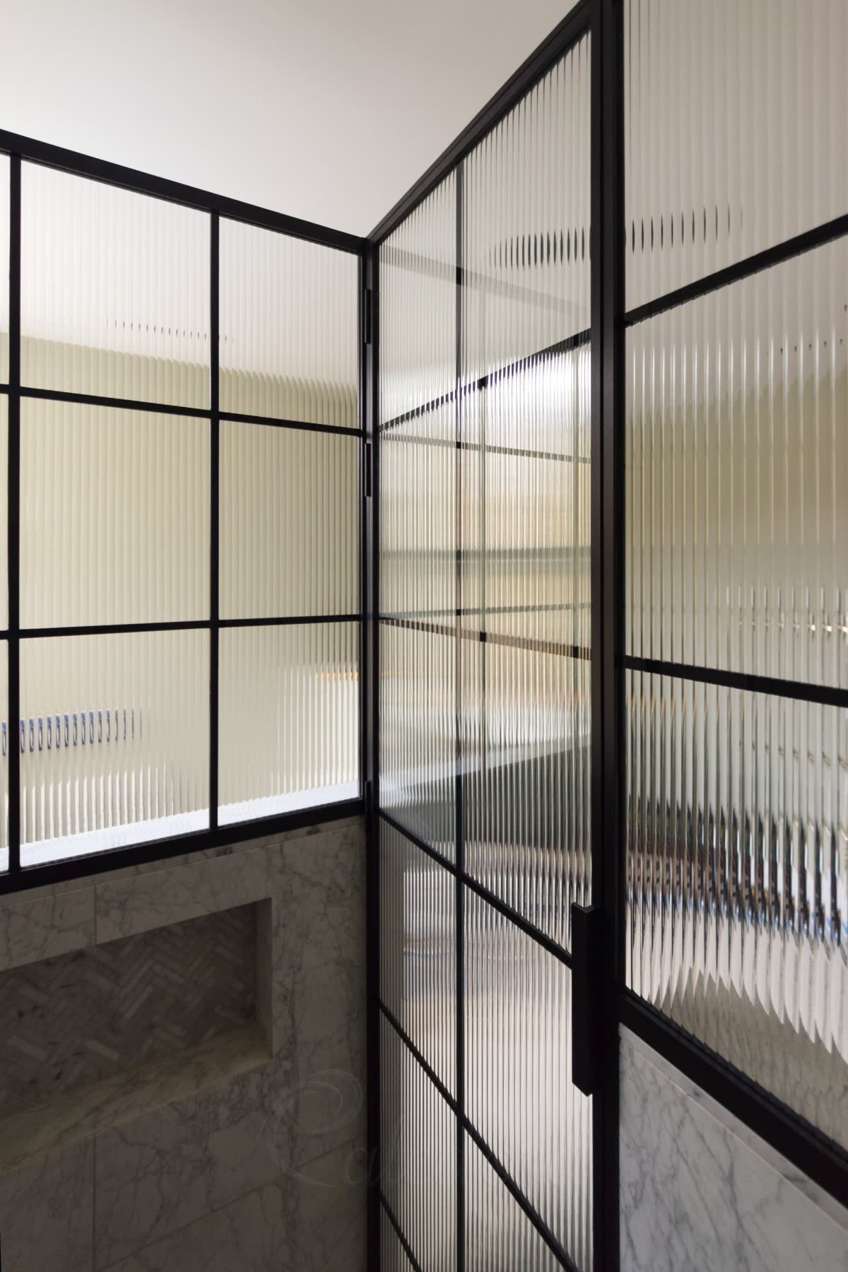 Textured glass shower enclosure photographed from inside — the vertical ribs sit on the outer face while broad unbroken reflections sweep across the flat inner face, in a black grid frame above marble