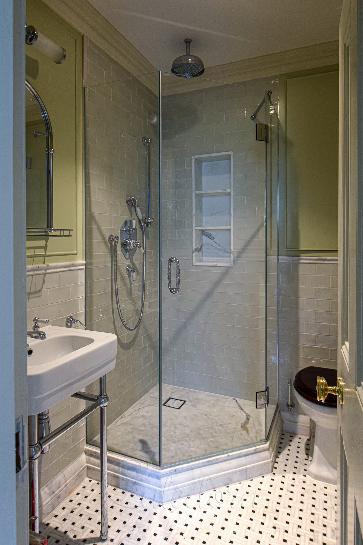 Frameless pentagonal shower enclosure with chamfered corner and angled door in period green-tile bathroom with marble tray and patterned floor