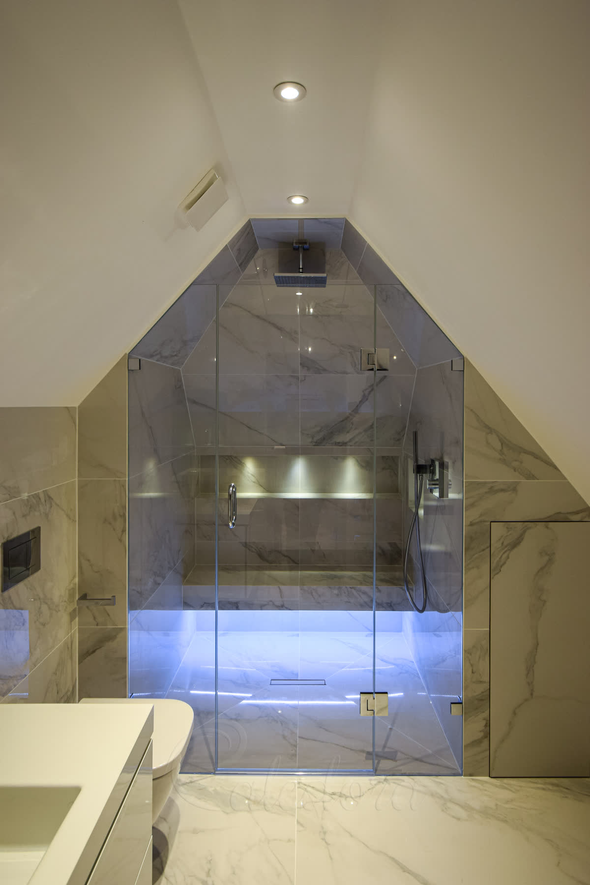 Frameless glass shower enclosure shaped to a sloped loft ceiling, with book-matched marble walls, built-in bench seating, chrome shower fixtures and LED floor strip in a London bathroom