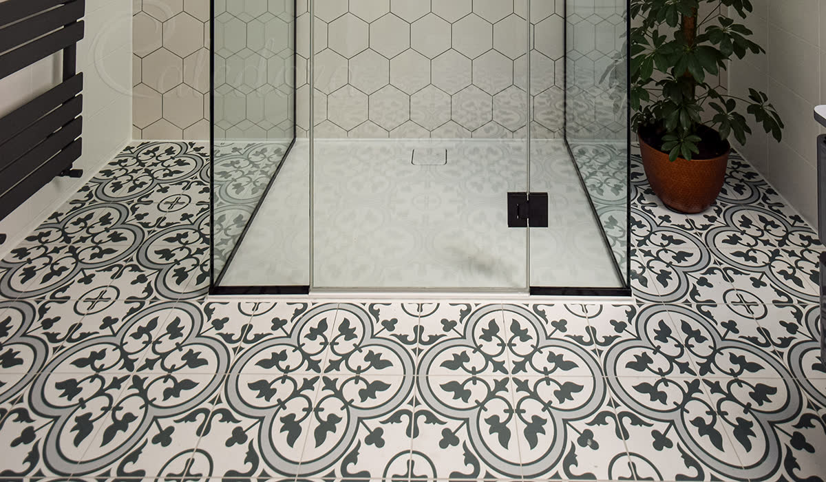 Frameless three-sided shower enclosure under a sloped loft ceiling with hexagon white tile walls, black fixtures and decorative patterned encaustic floor tiles