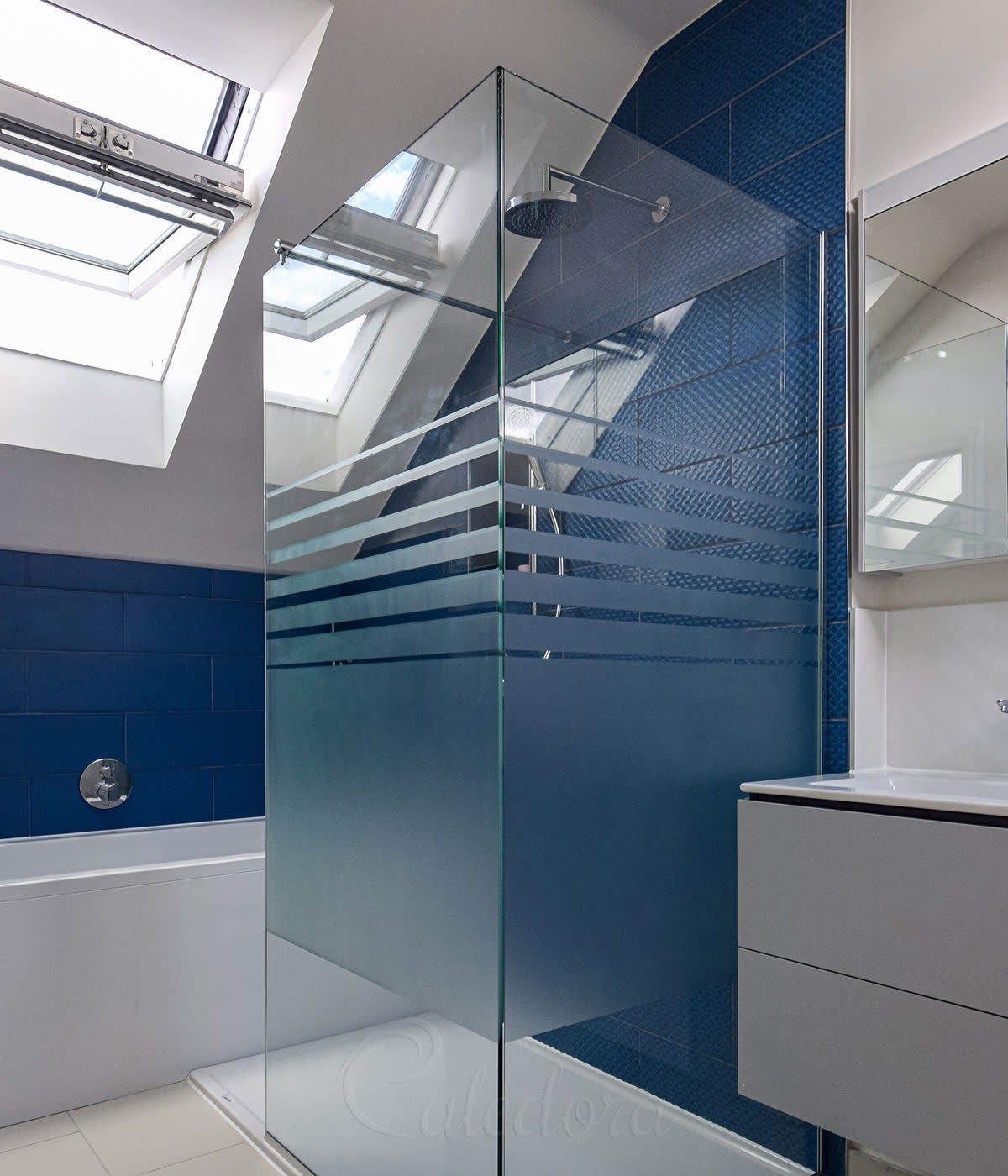 Frameless walk-in glass shower screen with horizontal sandblasted privacy bands, two-panel corner setup shaped to a sloped loft ceiling, in a London loft bathroom with blue tiles and skylight