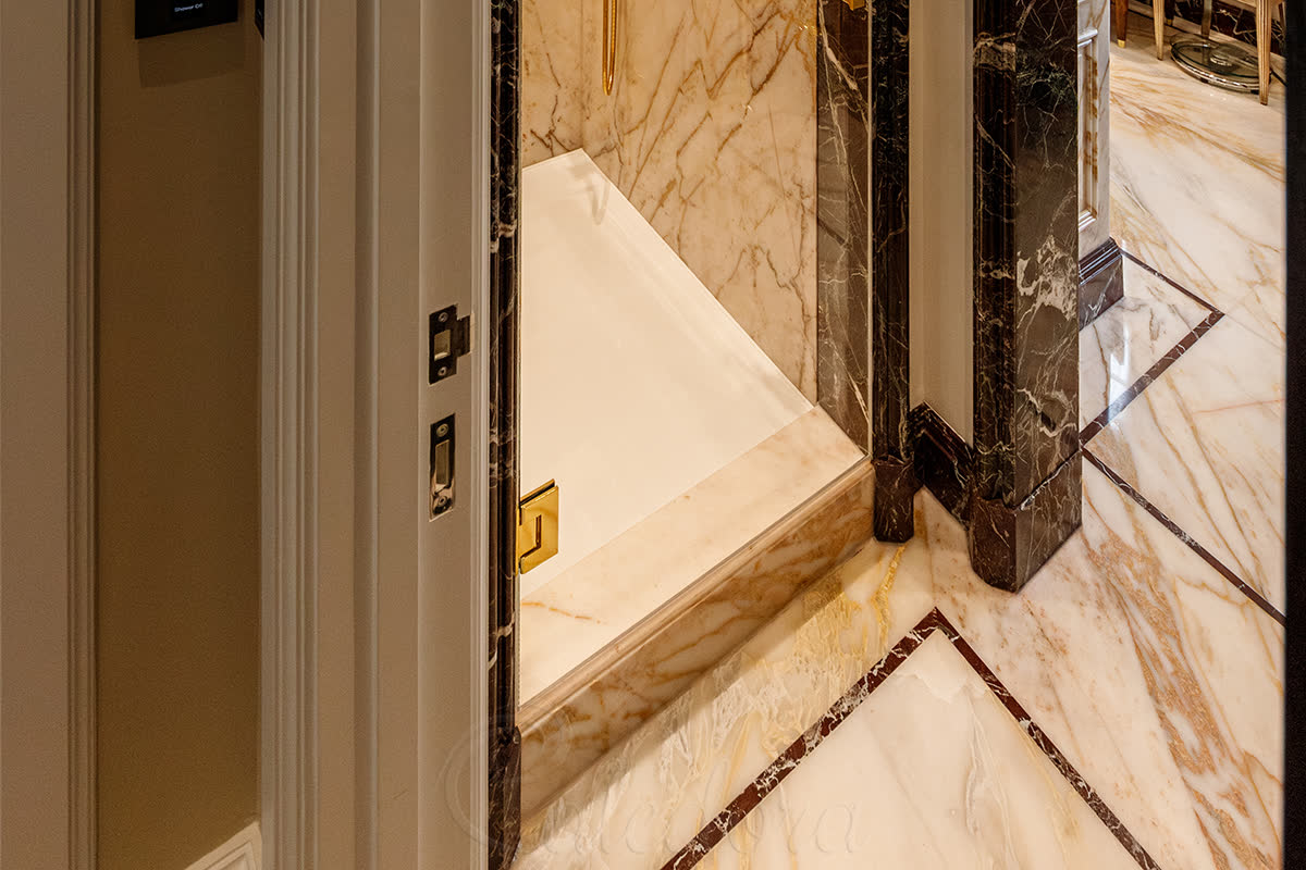 Frameless glass shower door with polished gold hardware closed against book-matched dark-veined marble walls with built-in marble bench seat in a luxury London bathroom
