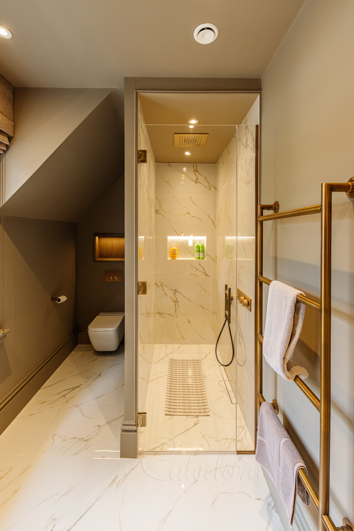 Frameless antique brass shower door in a Leicester wet room with marble walls, inline fixed panel held in antique brass U-channels and matching ceiling rain shower
