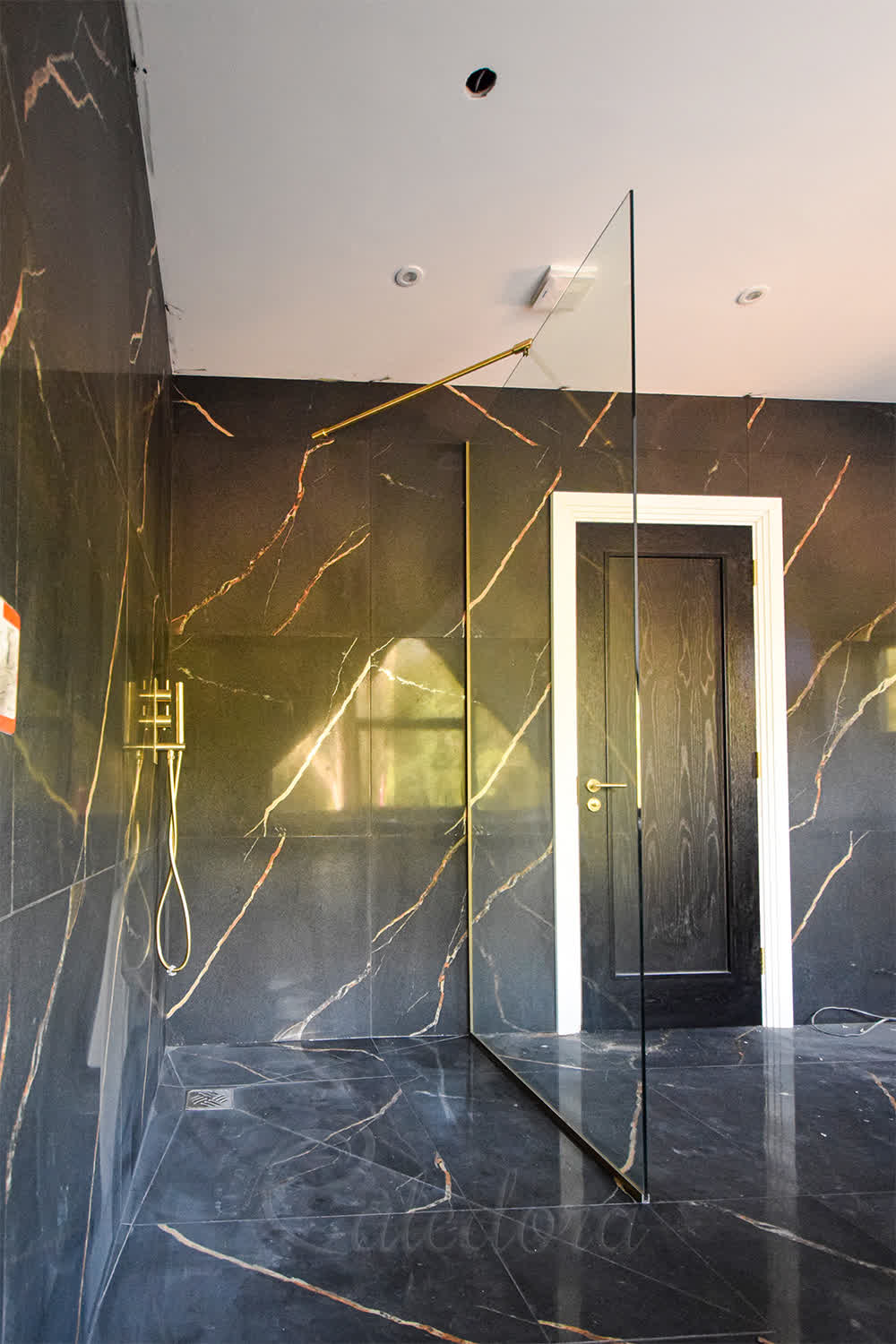 Walk-in shower screen with brushed gold hardware and 45-degree reinforcement bar in marble-effect bathroom during construction