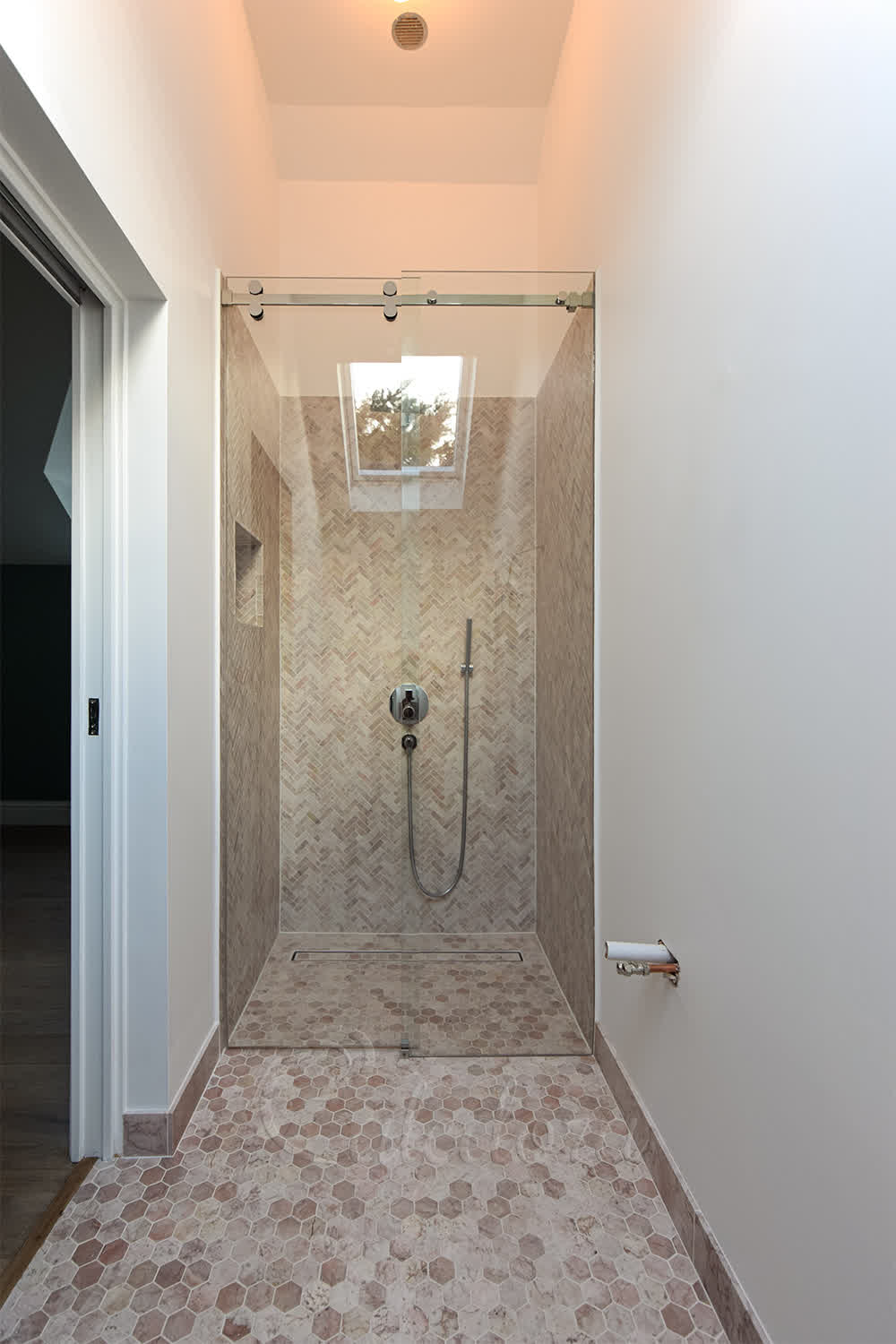 Frameless sliding shower door closed with polished chrome track and herringbone tile walls in refurbishment bathroom