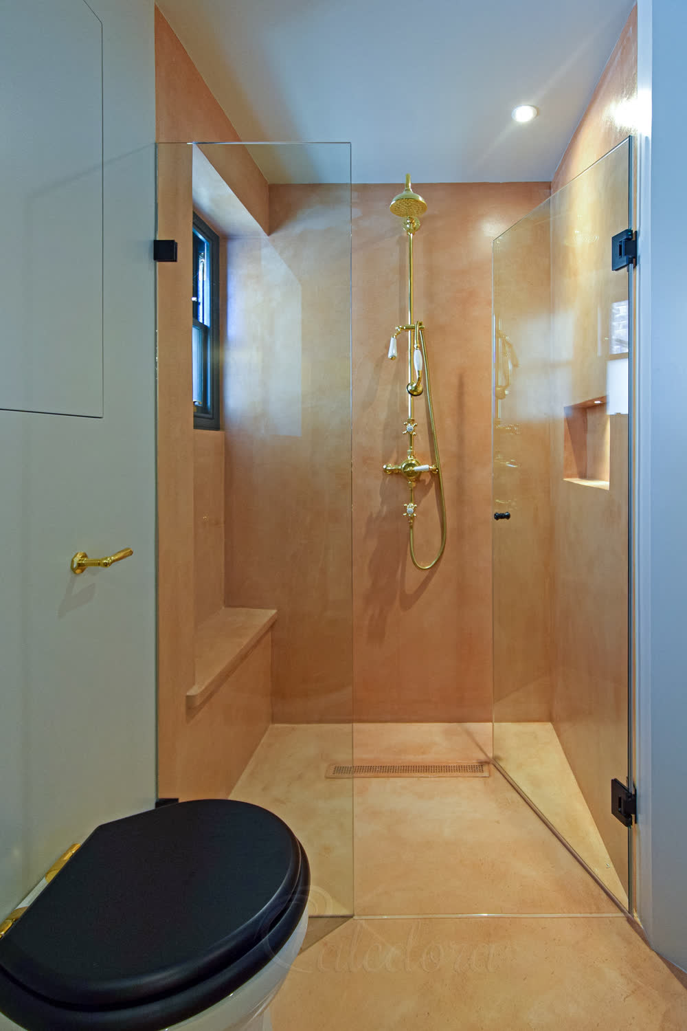 Frameless shower door with rectangular black hinges partially open next to an inline fixed panel, peach microcement walls and brass shower fixtures in a London bathroom