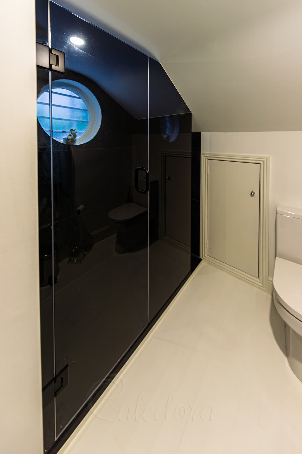 Dark grey tinted 10mm glass shower enclosure reading as near-solid black from outside, under a slanted ceiling with a round window in a London loft bathroom