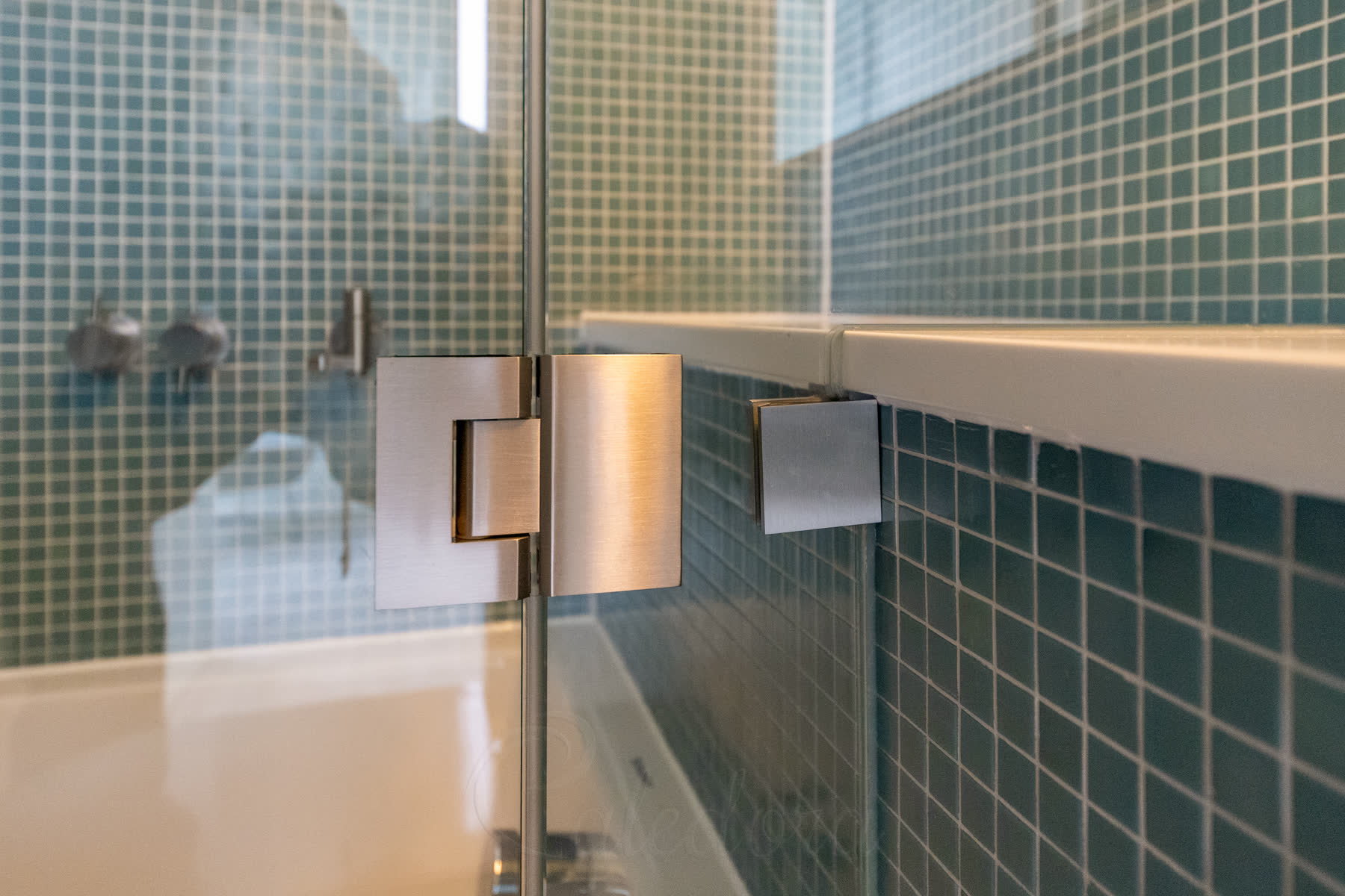Close-up of the brushed nickel bottom hinge between fixed panel and hinged bath panel, with wall bracket holding the fixed panel under the corian shelf