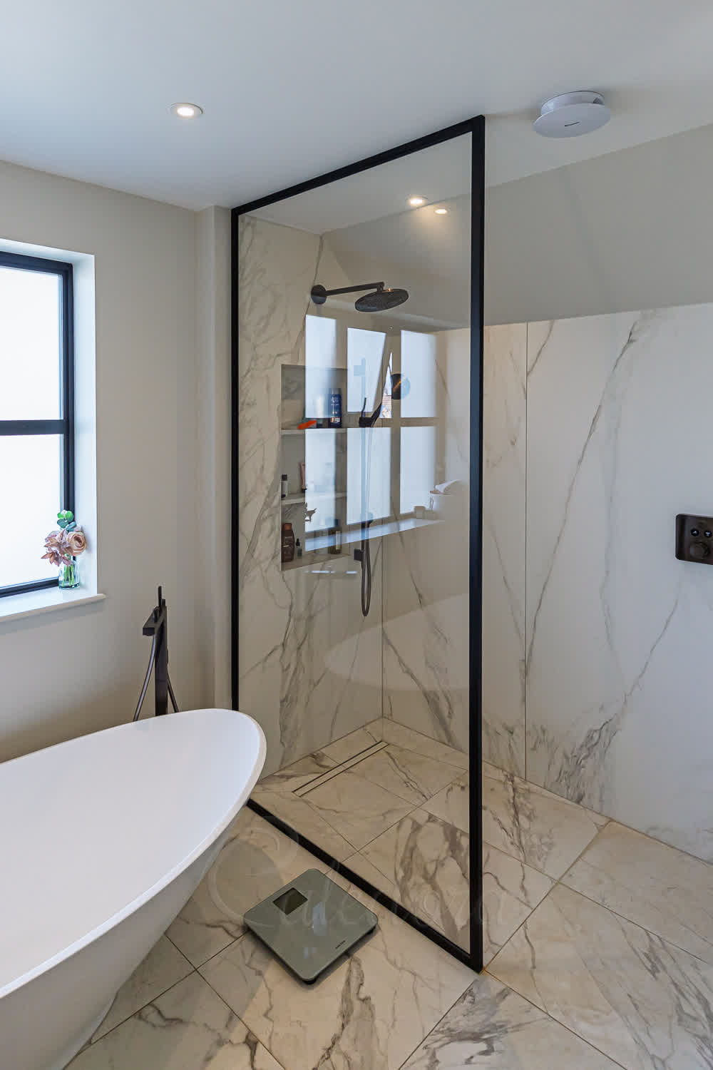 Matt black framed walk-in shower screen with marble-effect tiles and white freestanding bath in modern bathroom