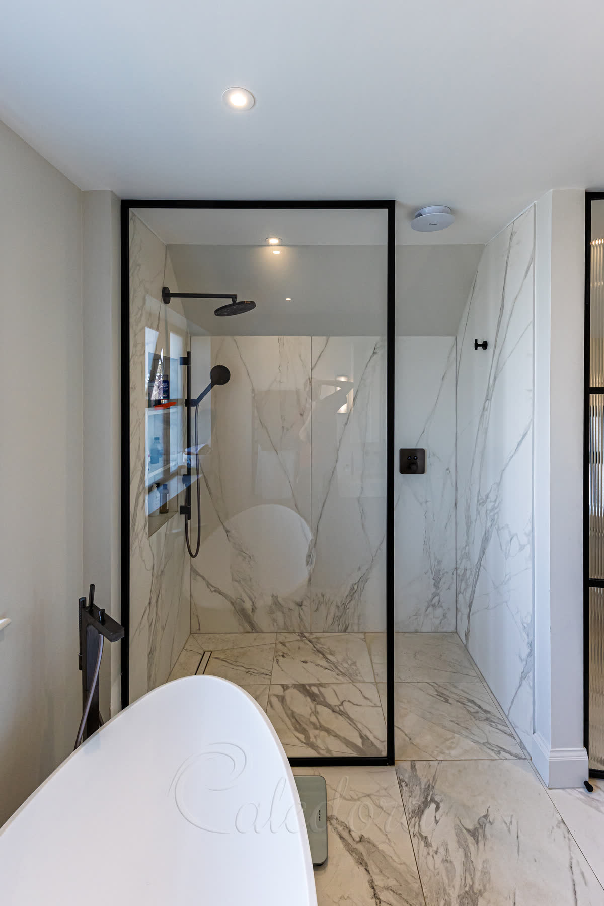 Matt black framed walk-in shower screen with marble-effect tiles and white freestanding bath in modern bathroom
