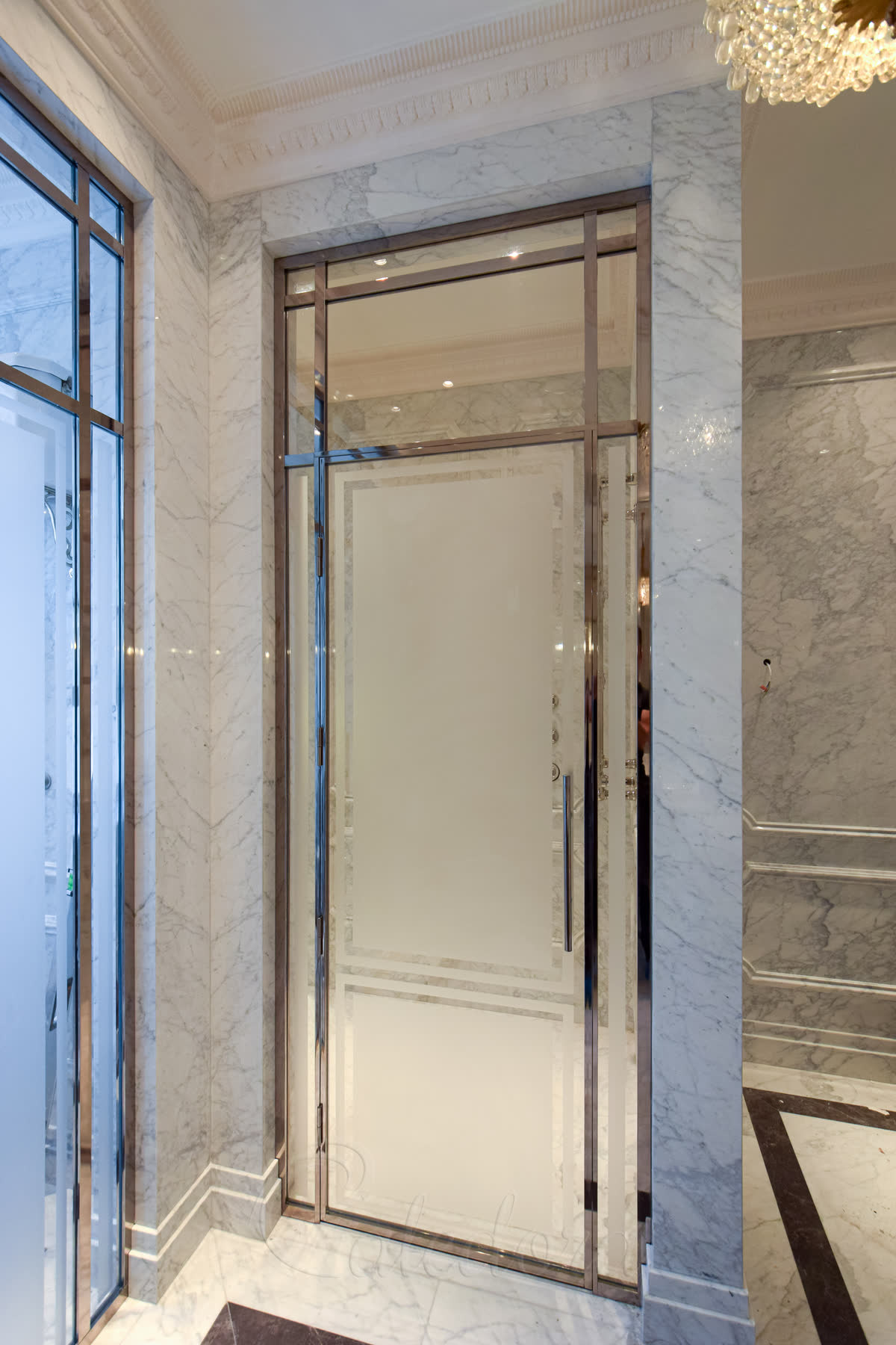 Closed nickel shower door with elegant mirror-polished frame against marble walls and crystal chandelier